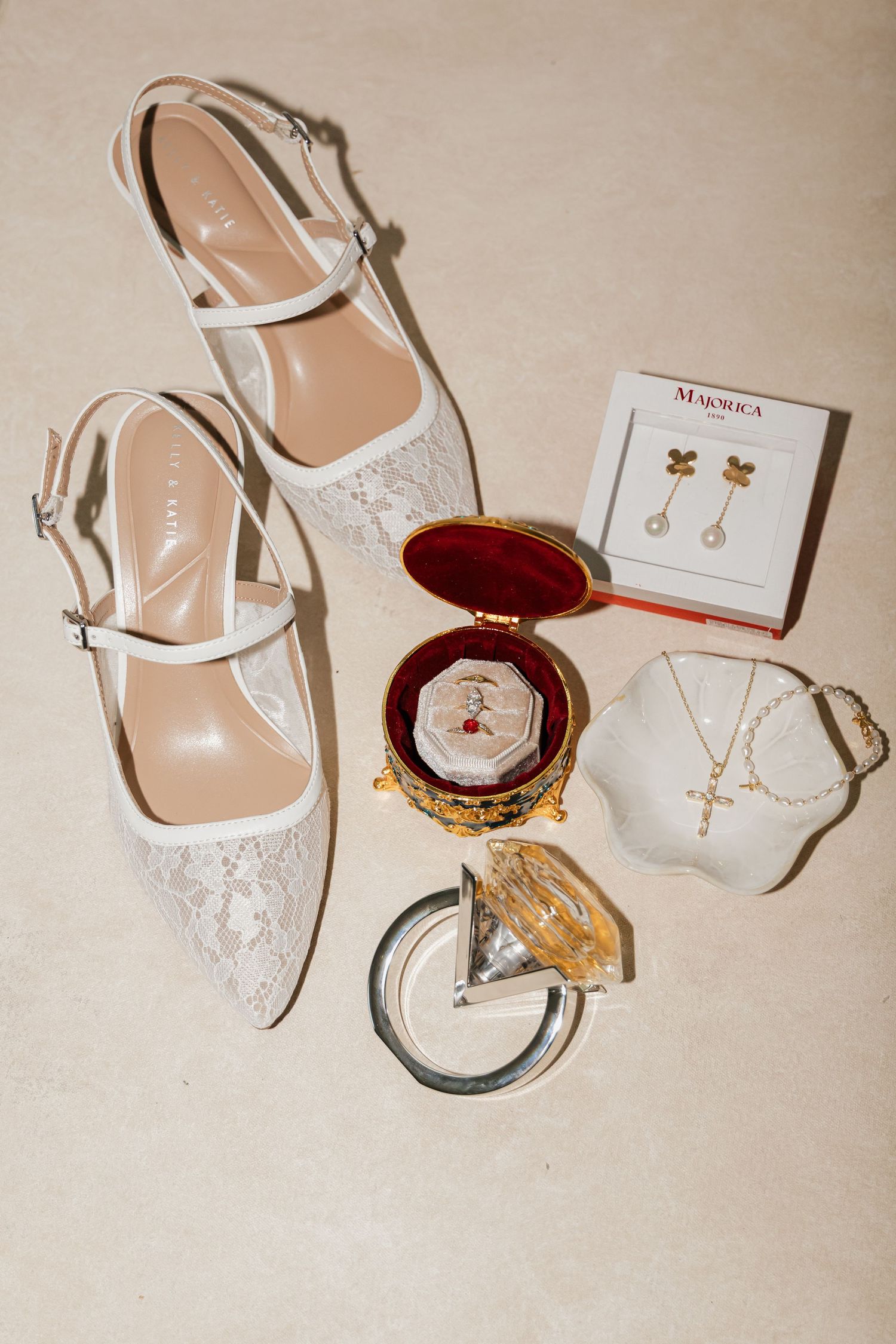 White lace bridal heels displayed with gold earrings, cross necklace, and ornate jewelry box on neutral background.