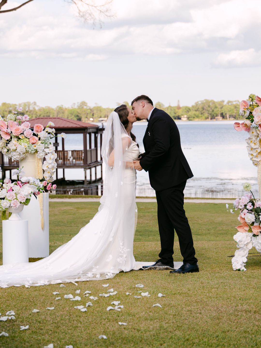Bride and groom share first kiss at lakeside altar surrounded by pink and white floral arrangements.