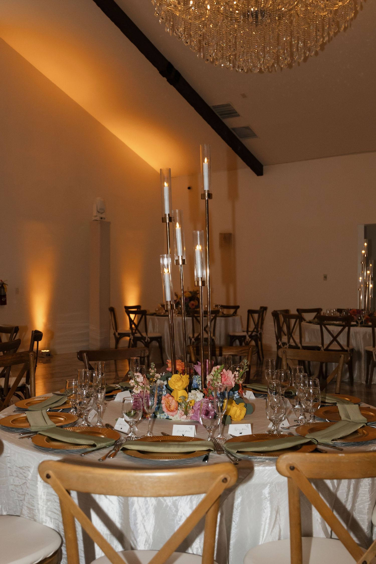 Elegant wedding reception table with tall candelabra centerpiece, colorful florals, and crystal chandelier overhead.