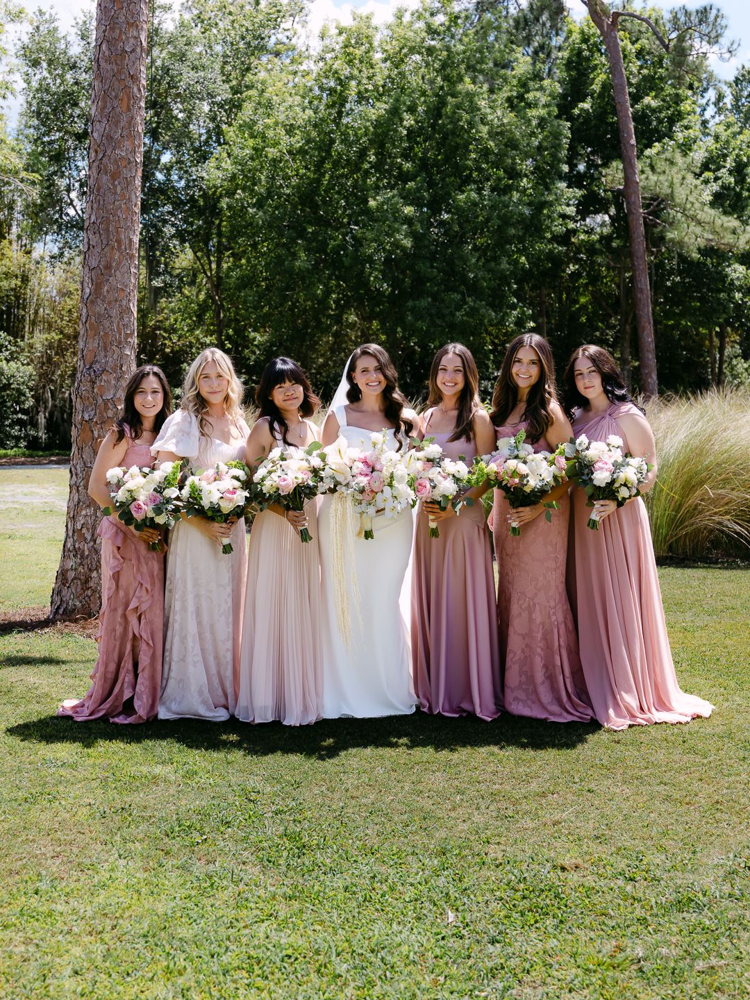 Bride with six bridesmaids in pink dresses holding floral bouquets outdoors.