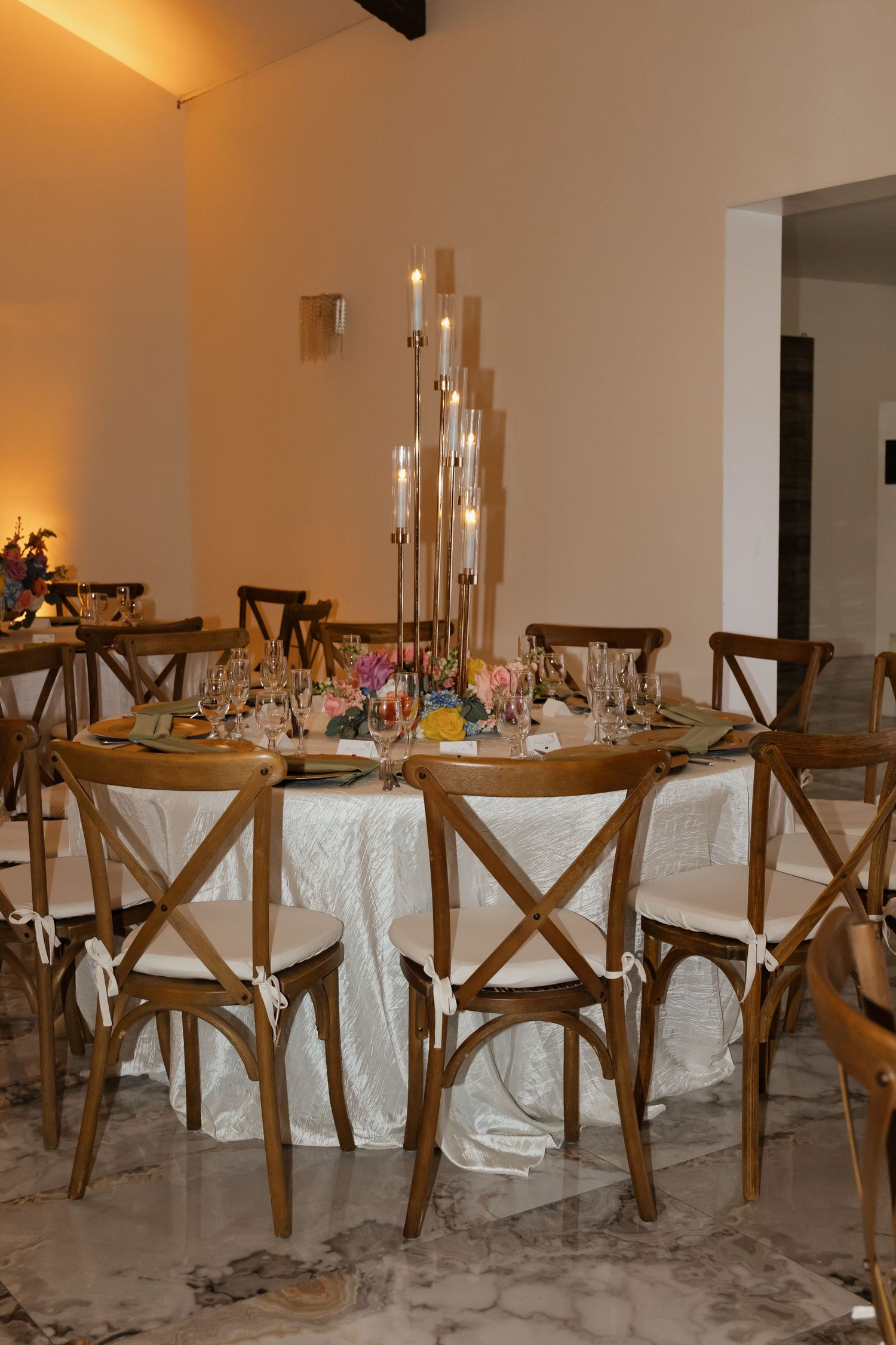 Romantic wedding reception table with wooden cross-back chairs and tall gold candelabra centerpiece.