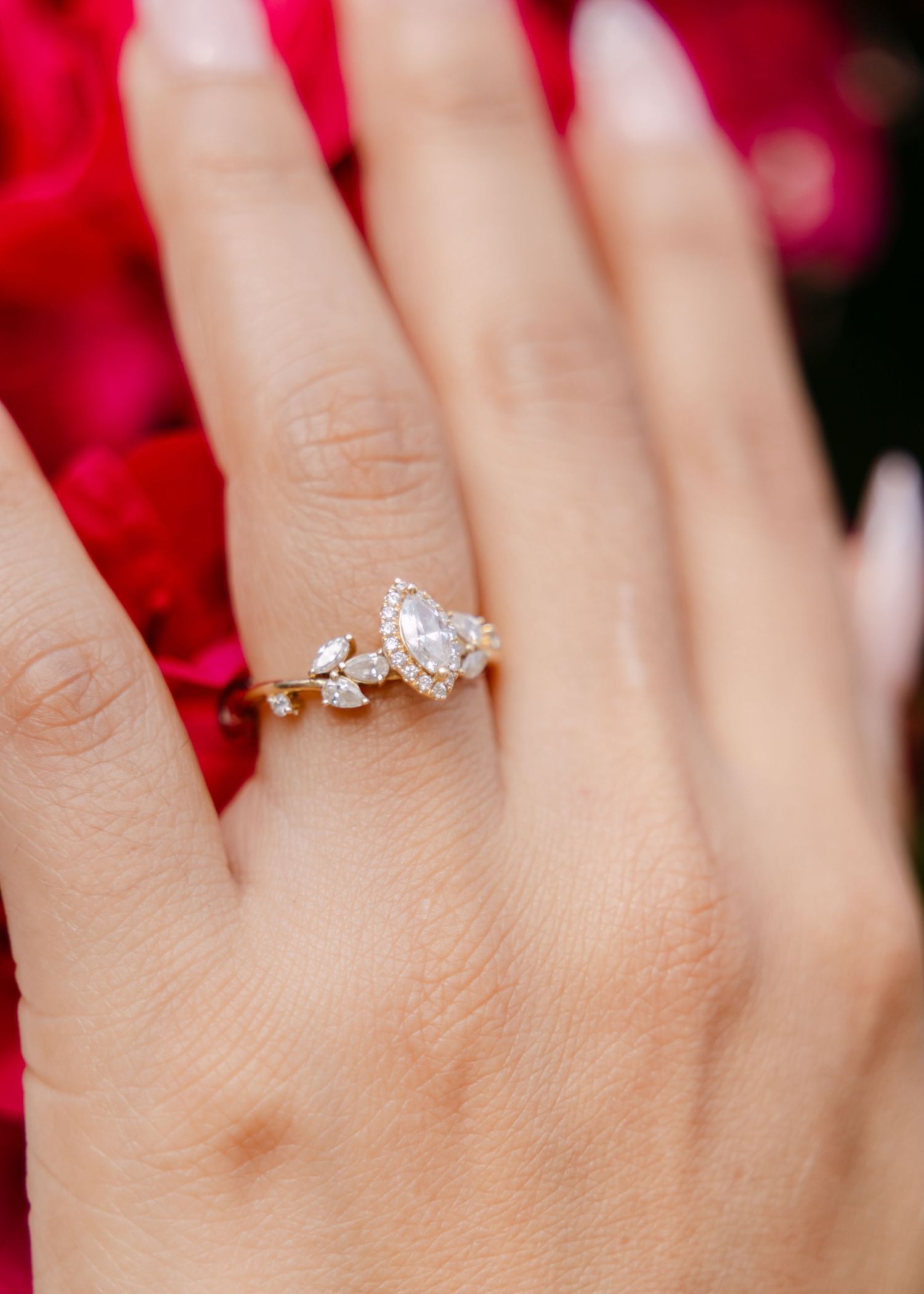 Close-up of a gold pear-shaped diamond engagement ring with cluster accents on a hand near red roses.