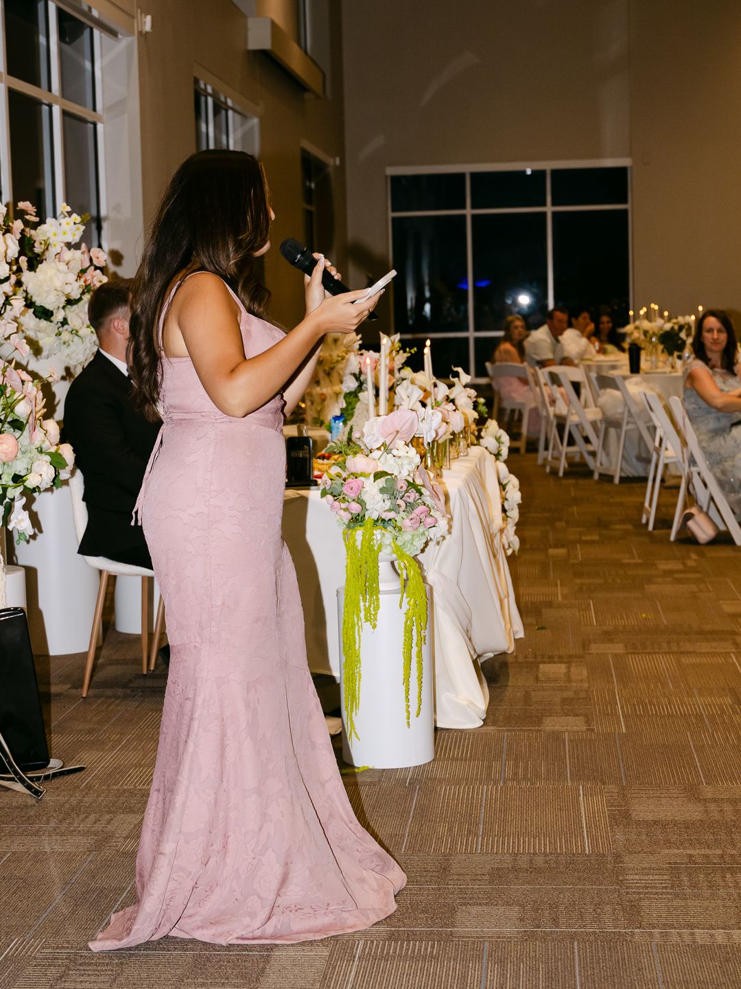 Bridesmaid in pink gown giving speech at elegant wedding reception with floral arrangements and candlelit tables.