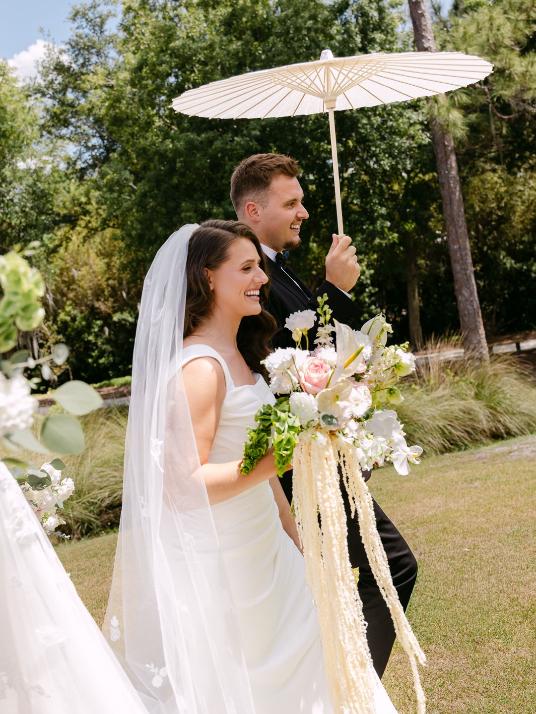 Bride and groom walk outdoors holding a white parasol, bride carrying a floral bouquet with trailing ribbons on a sunny day.