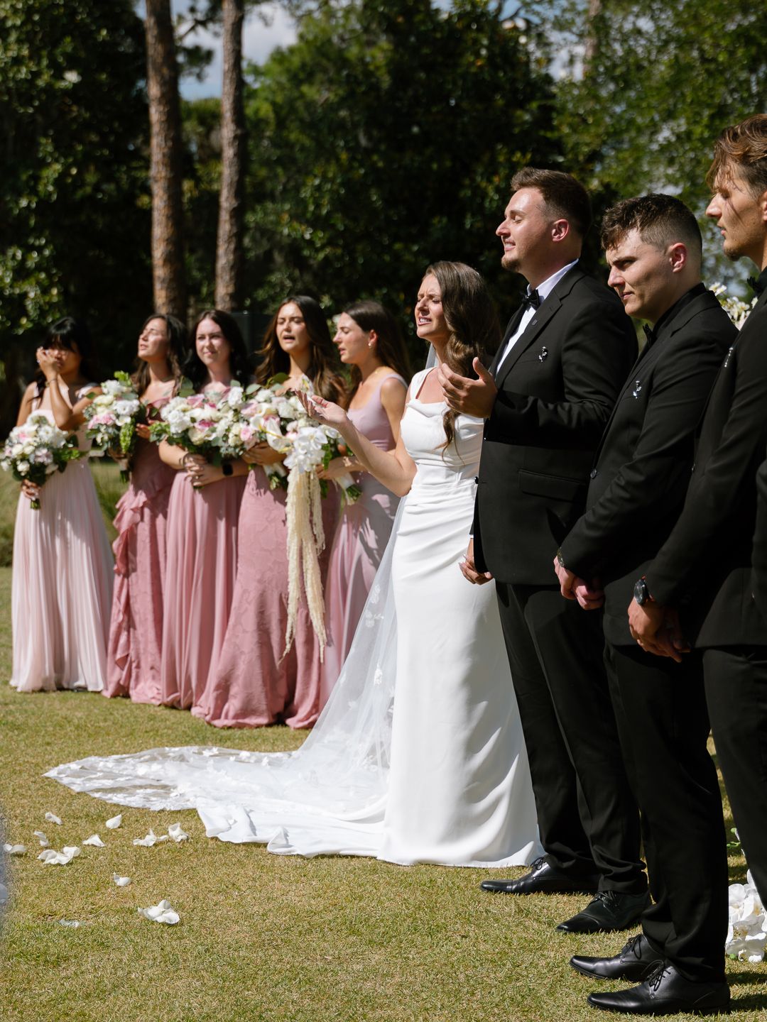 Wedding party in pink bridesmaid dresses and black tuxedos stand together at outdoor ceremony.