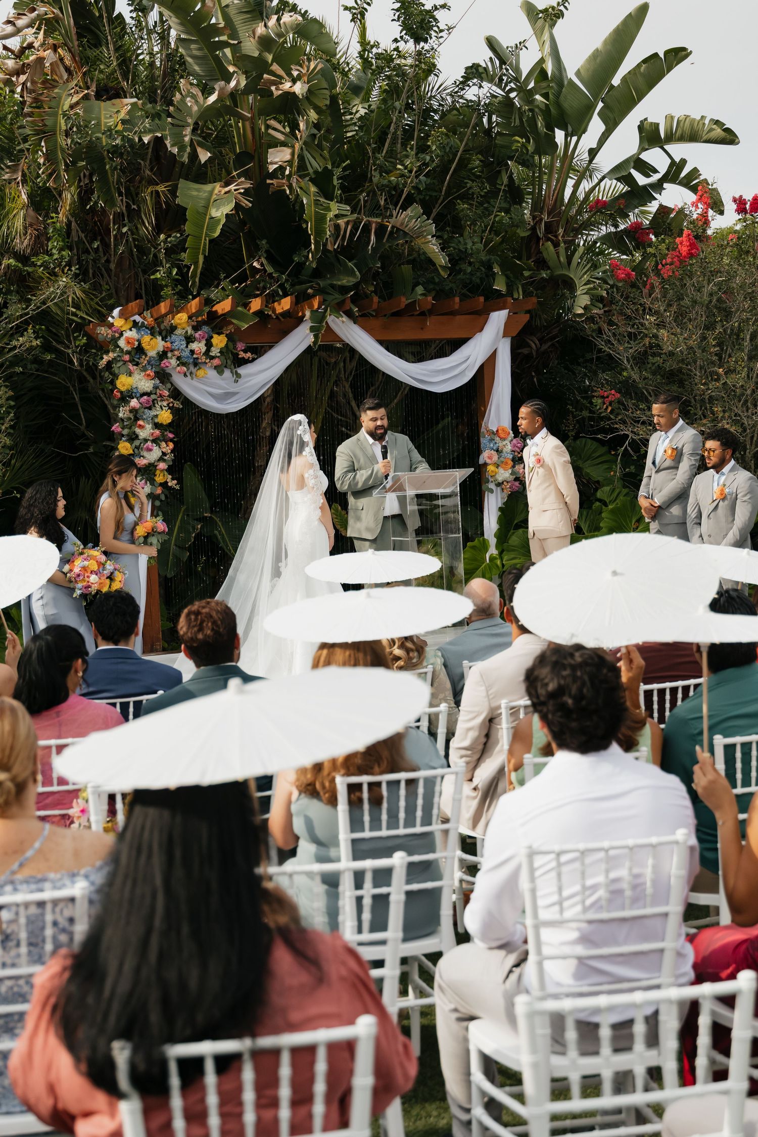 Outdoor wedding ceremony with guests holding white parasols seated in white chairs before a floral arch altar.