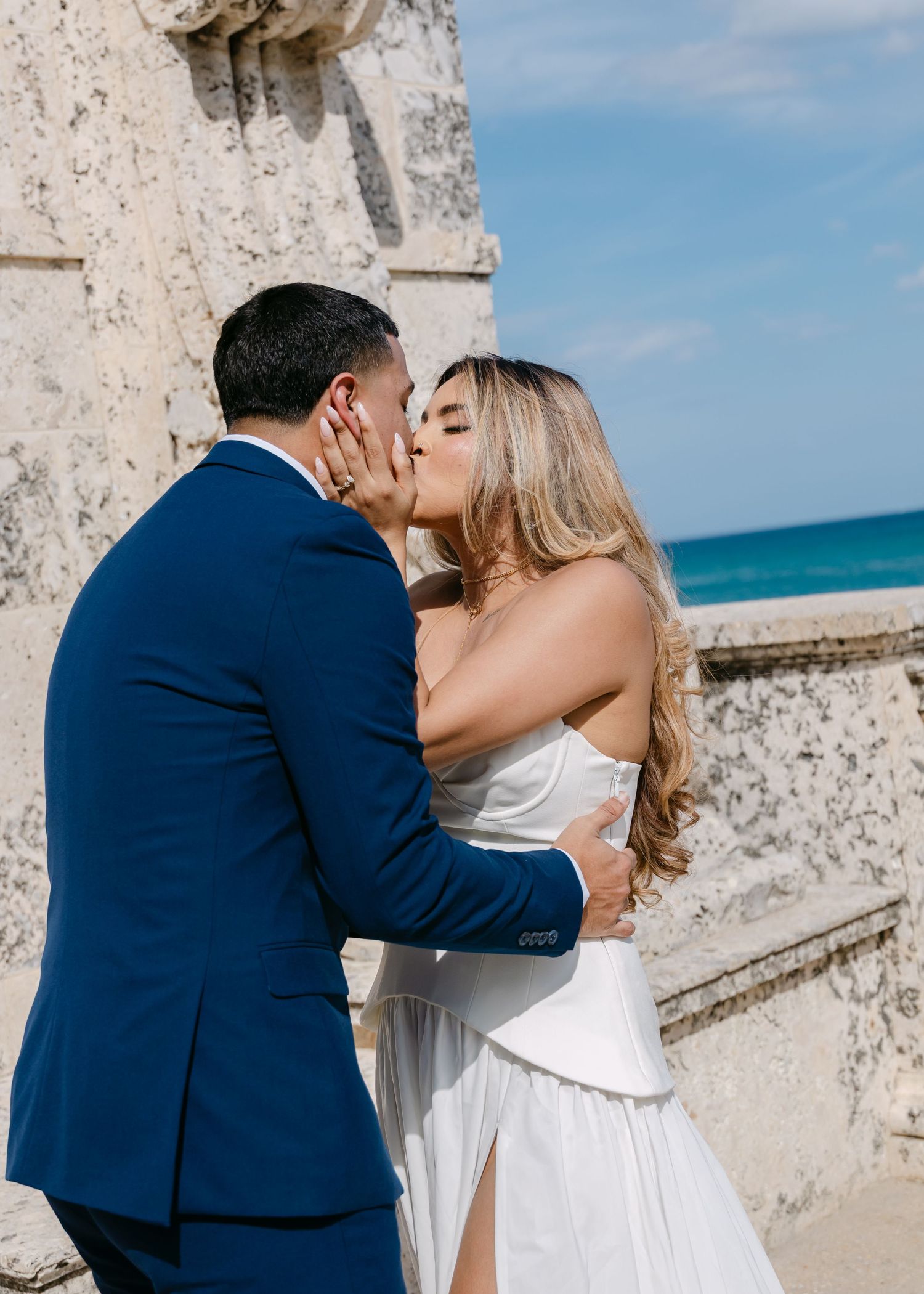 Couple sharing a romantic kiss in wedding attire by a stone wall with ocean views.