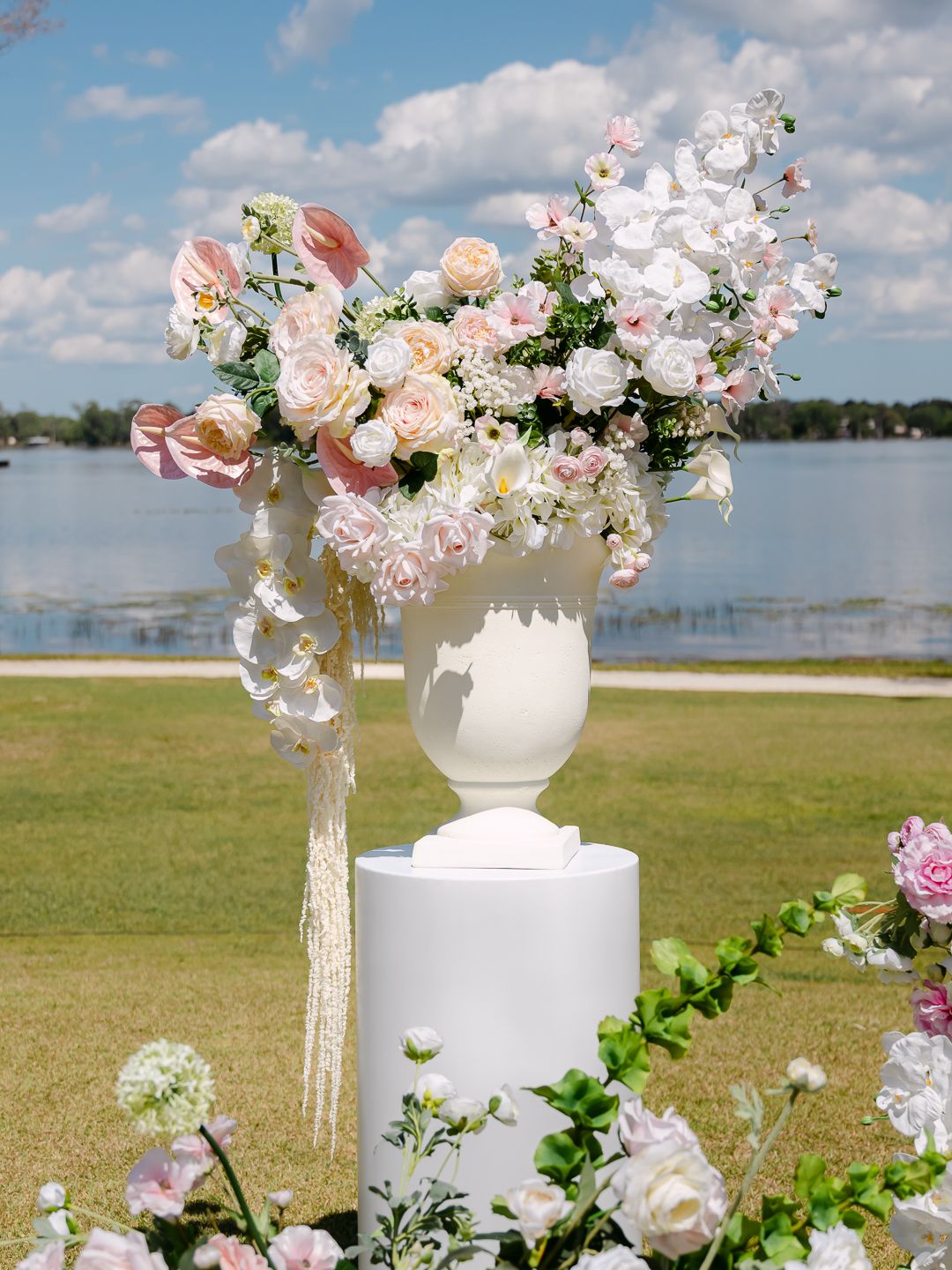 Elegant white urn filled with pink roses and white orchids on pedestal at lakeside wedding ceremony.