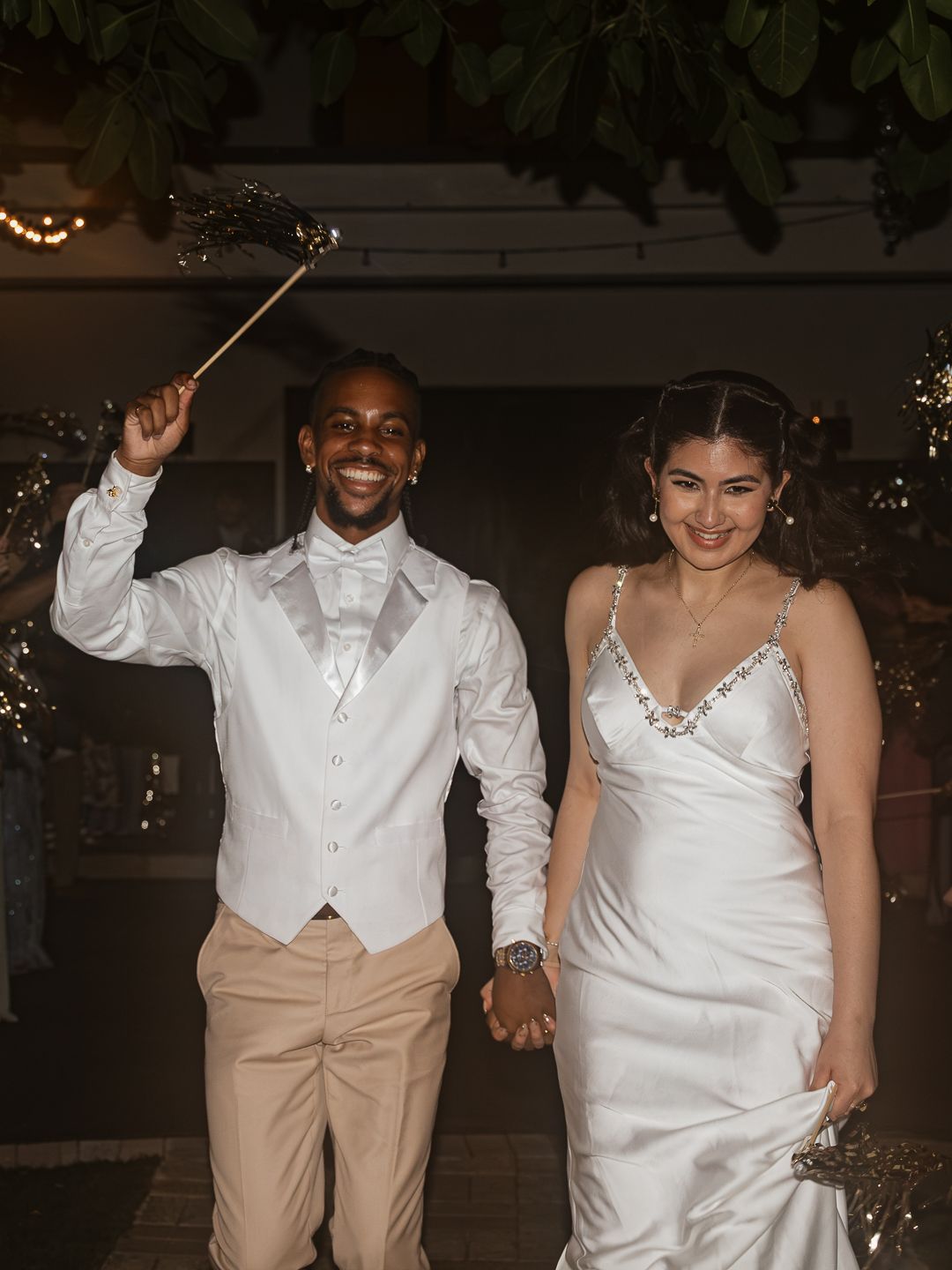 Smiling newlyweds exit wedding venue hand in hand as groom raises sparkler in celebration at night.