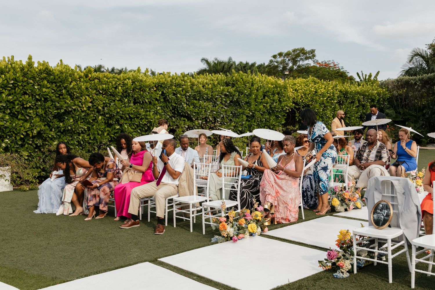 Wedding guests seated in white chairs holding parasols at an outdoor garden ceremony on a cloudy day.