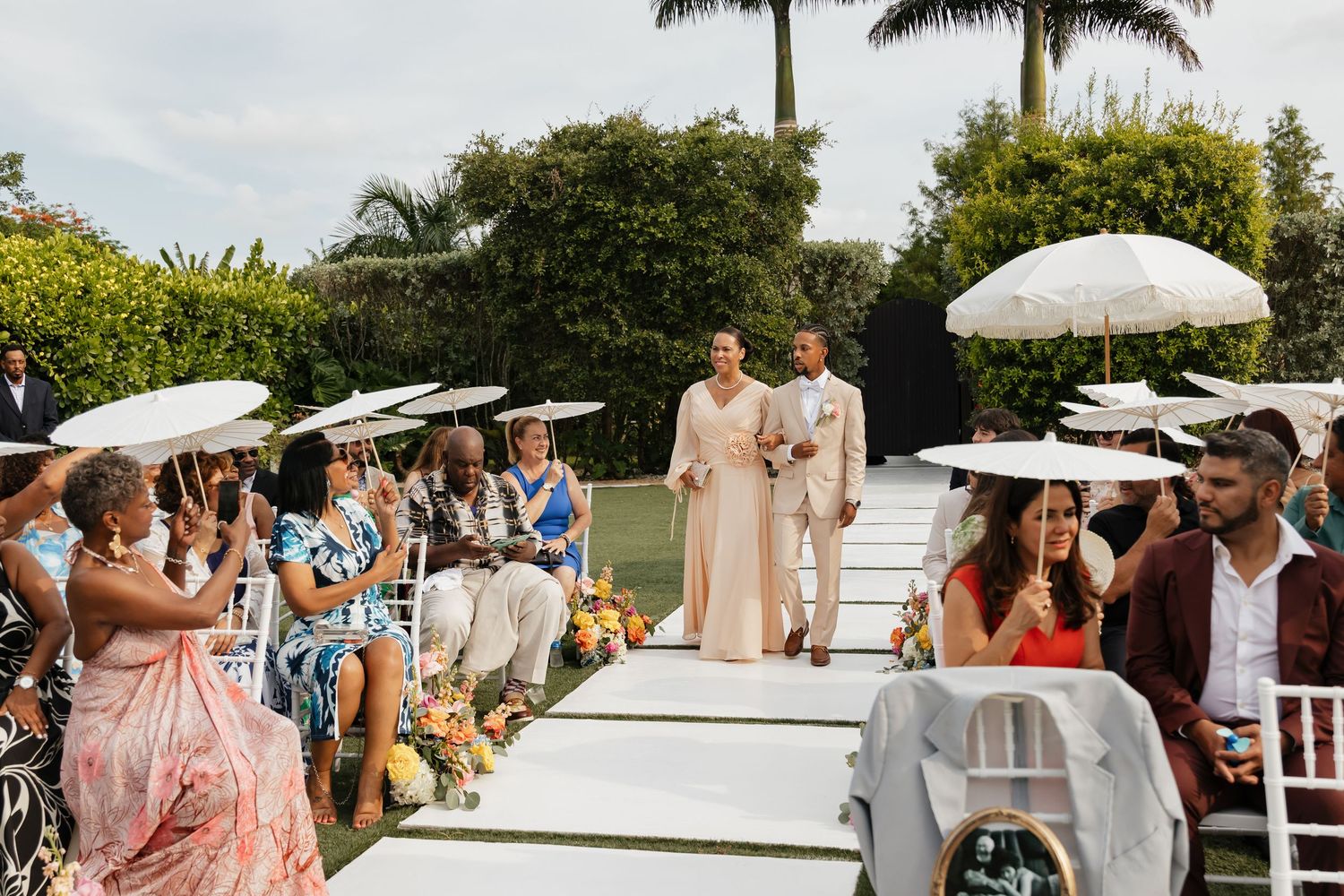 Bride and groom walk down white aisle together at outdoor garden wedding as guests watch and celebrate.