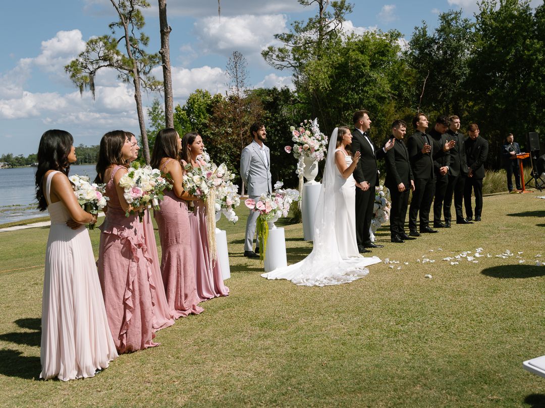 Wedding party standing at outdoor lakeside ceremony with bridesmaids in pink dresses and groomsmen in black suits.