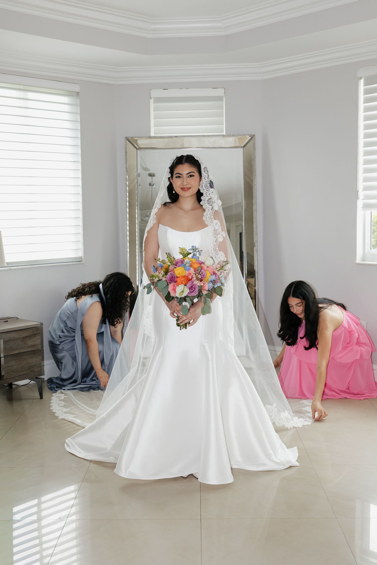 Bride in white satin gown holds colorful bouquet as bridesmaids in blue and pink adjust her train.