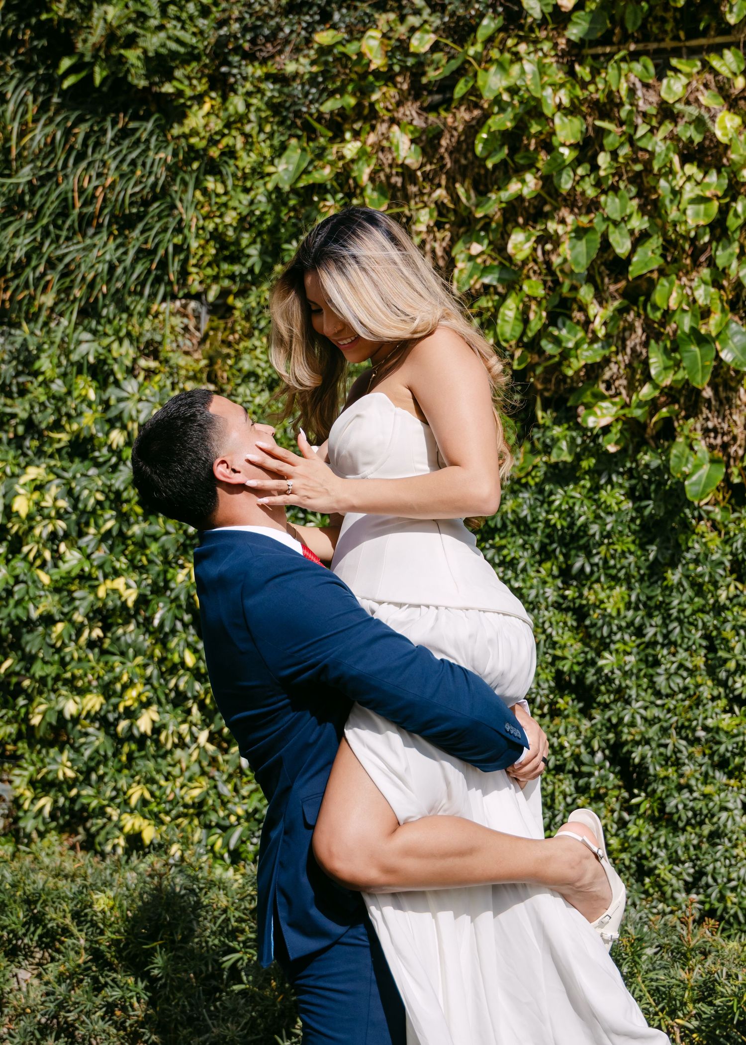 Groom in navy suit lifts bride in white wedding dress, sharing a kiss outdoors with lush green ivy backdrop.