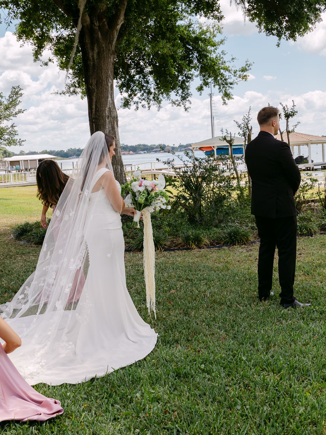 Bride in white gown and veil holds bouquet during outdoor first look near lakeside wedding venue.
