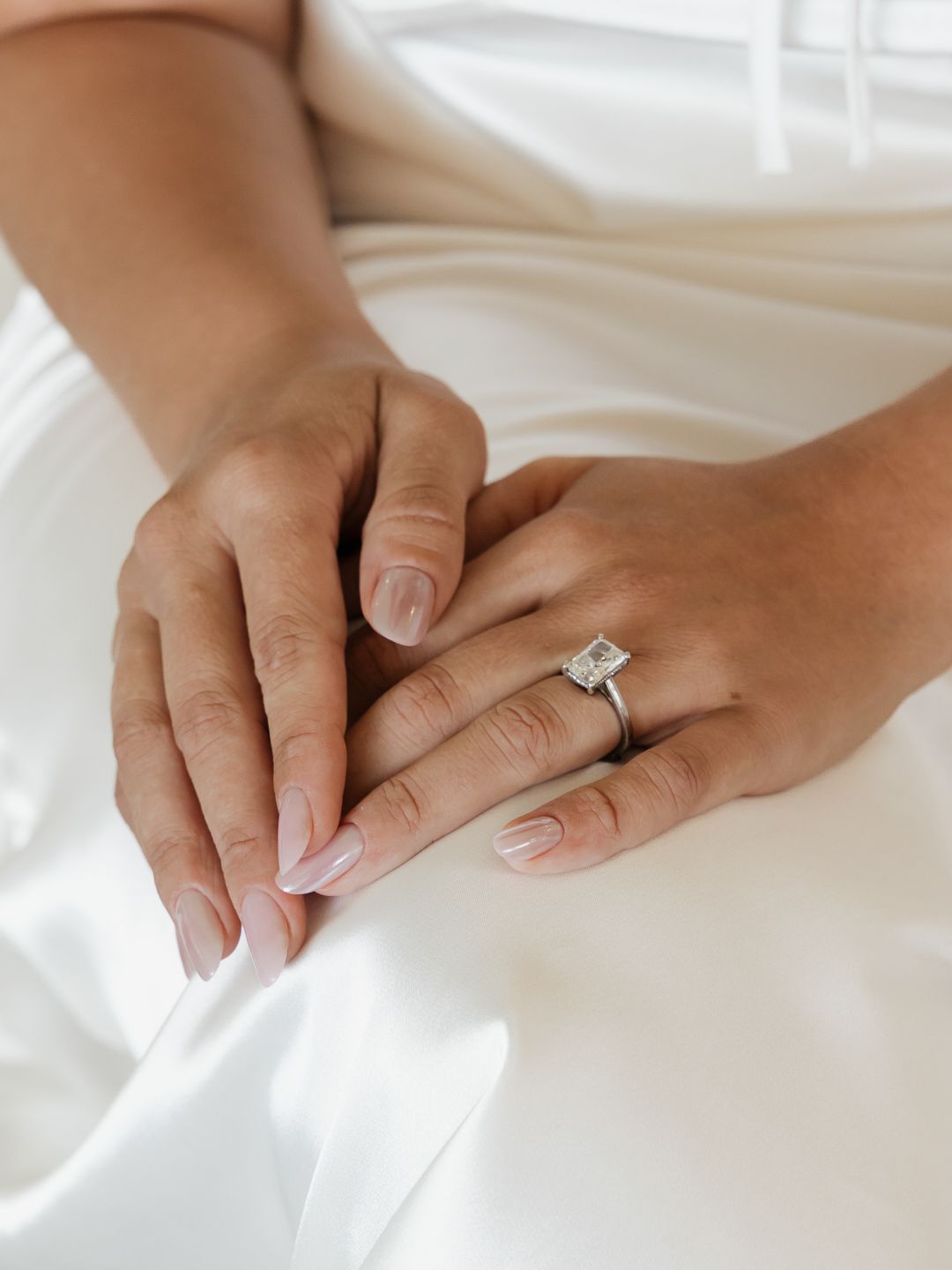 Close-up of bride's hands with elegant emerald-cut diamond engagement ring on white satin wedding dress.