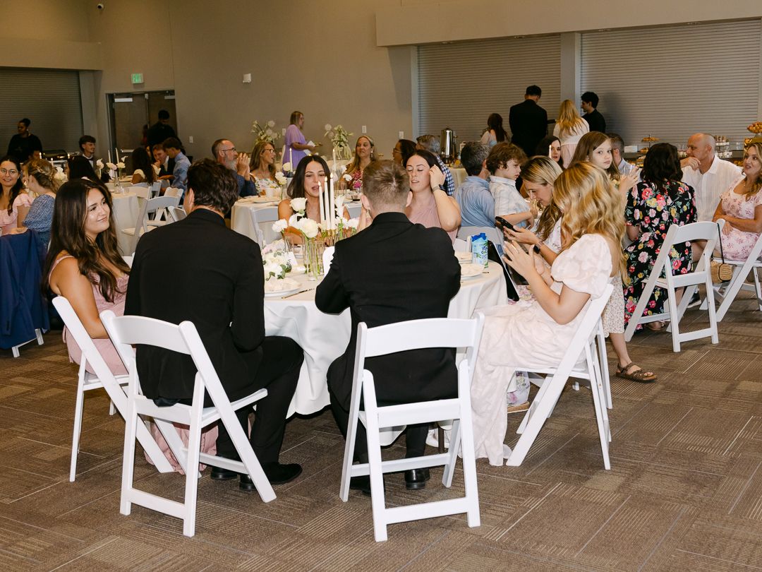 Wedding reception guests seated at round tables with white chairs in a bright banquet hall.
