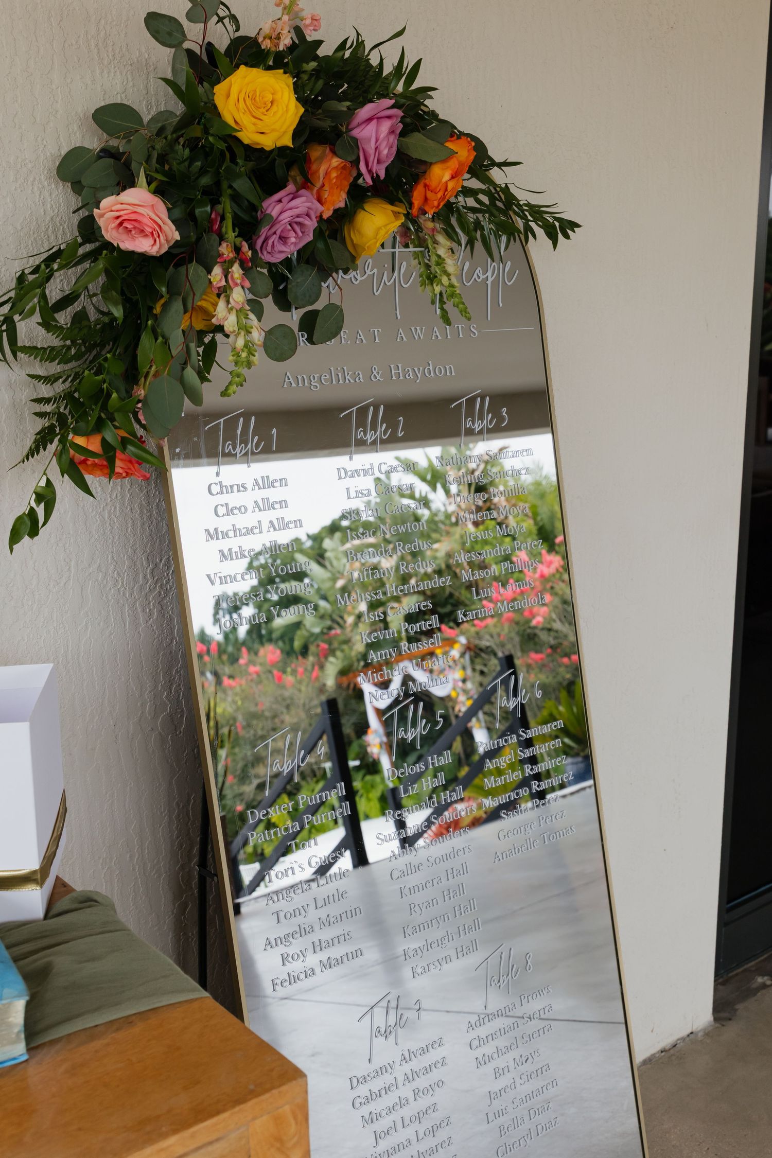 Wedding seating chart displayed on a mirror with colorful floral arrangement of yellow, pink, and orange roses on top.