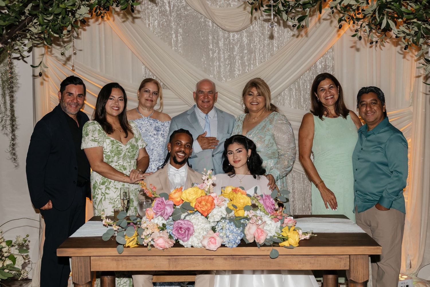 Wedding party group photo with bride and groom signing documents surrounded by family members.