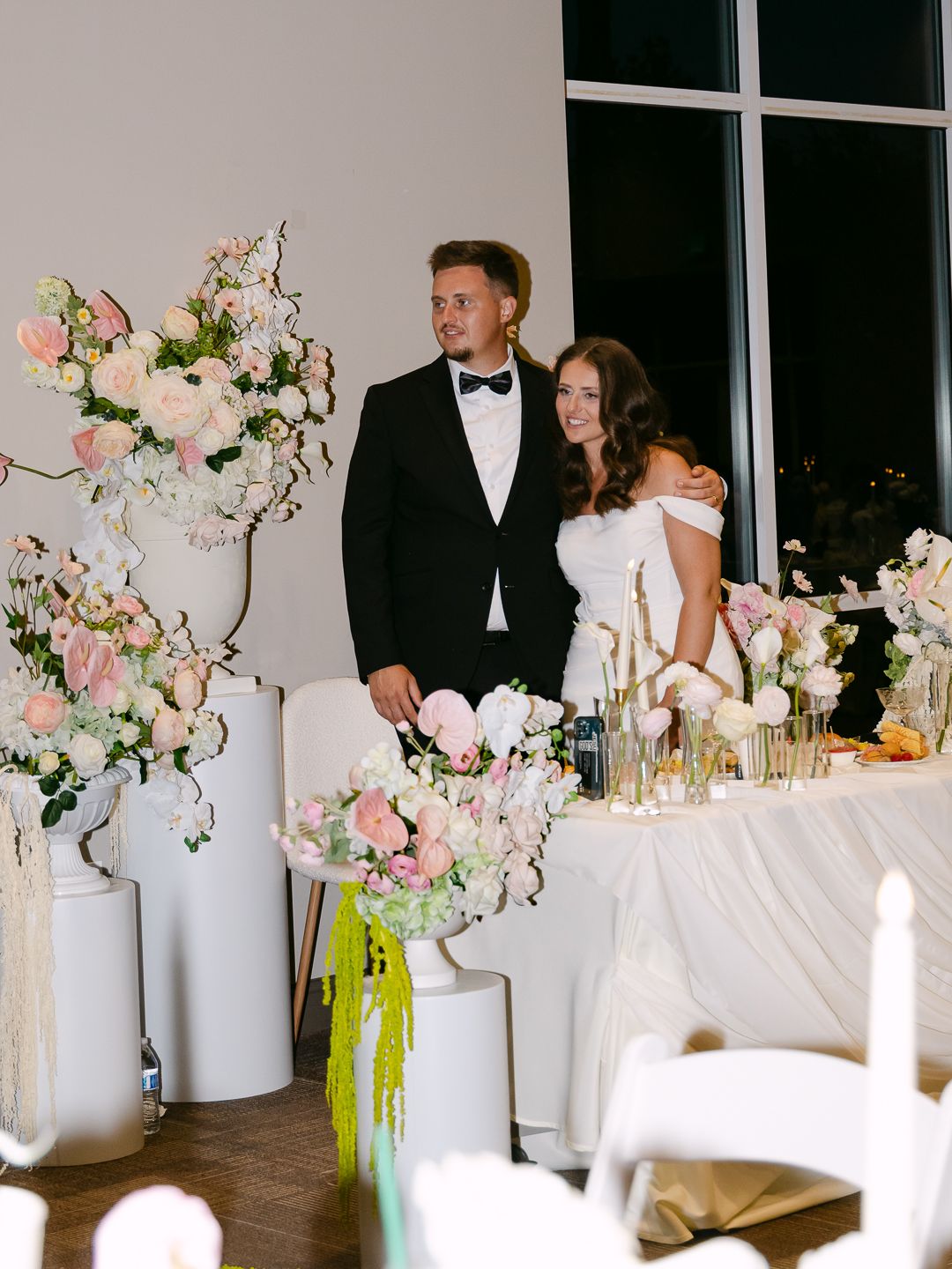 Bride and groom standing at sweetheart table surrounded by tall white floral arrangements and pink roses at reception.