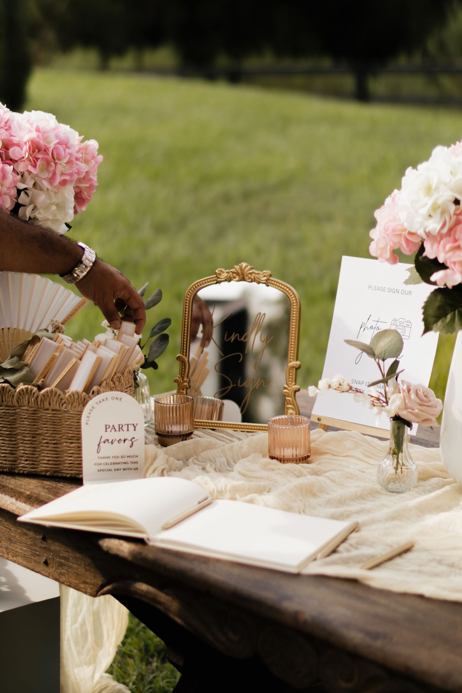 Elegant outdoor wedding welcome table with pink hydrangeas, party favors, guest book, and gold mirror decor on rustic wood.
