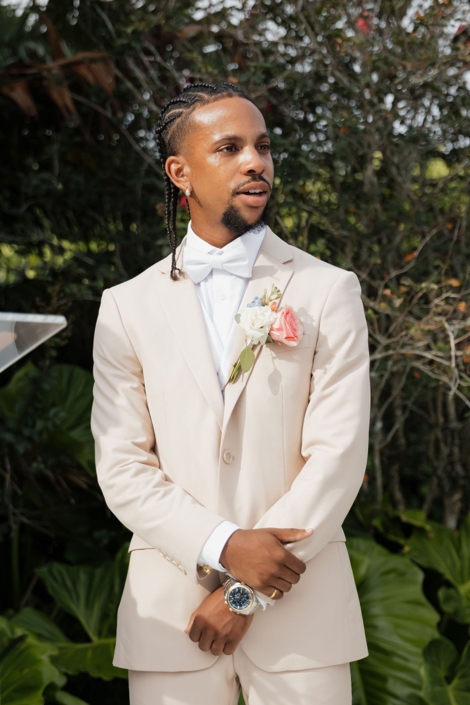 Groom in cream suit with floral boutonniere standing outdoors among tropical foliage.