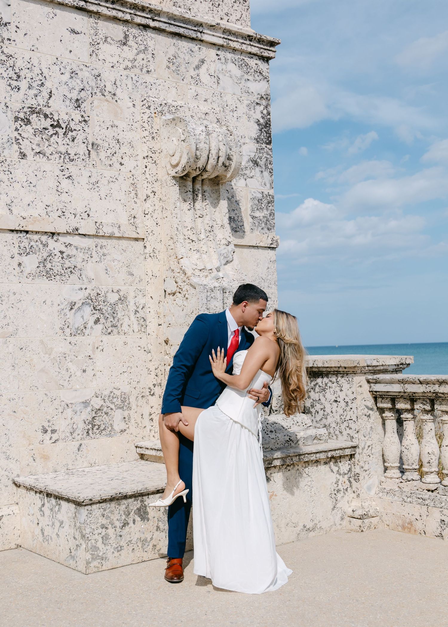 Couple shares romantic dip kiss in white wedding dress and navy suit by historic stone wall oceanside.