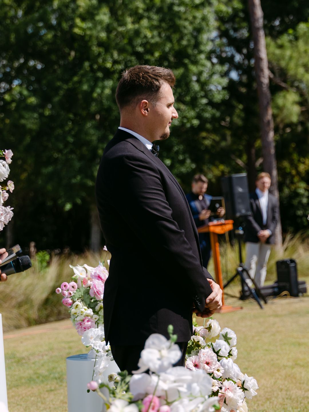 Groom in black tuxedo standing at outdoor wedding ceremony altar surrounded by pink and white floral arrangements.