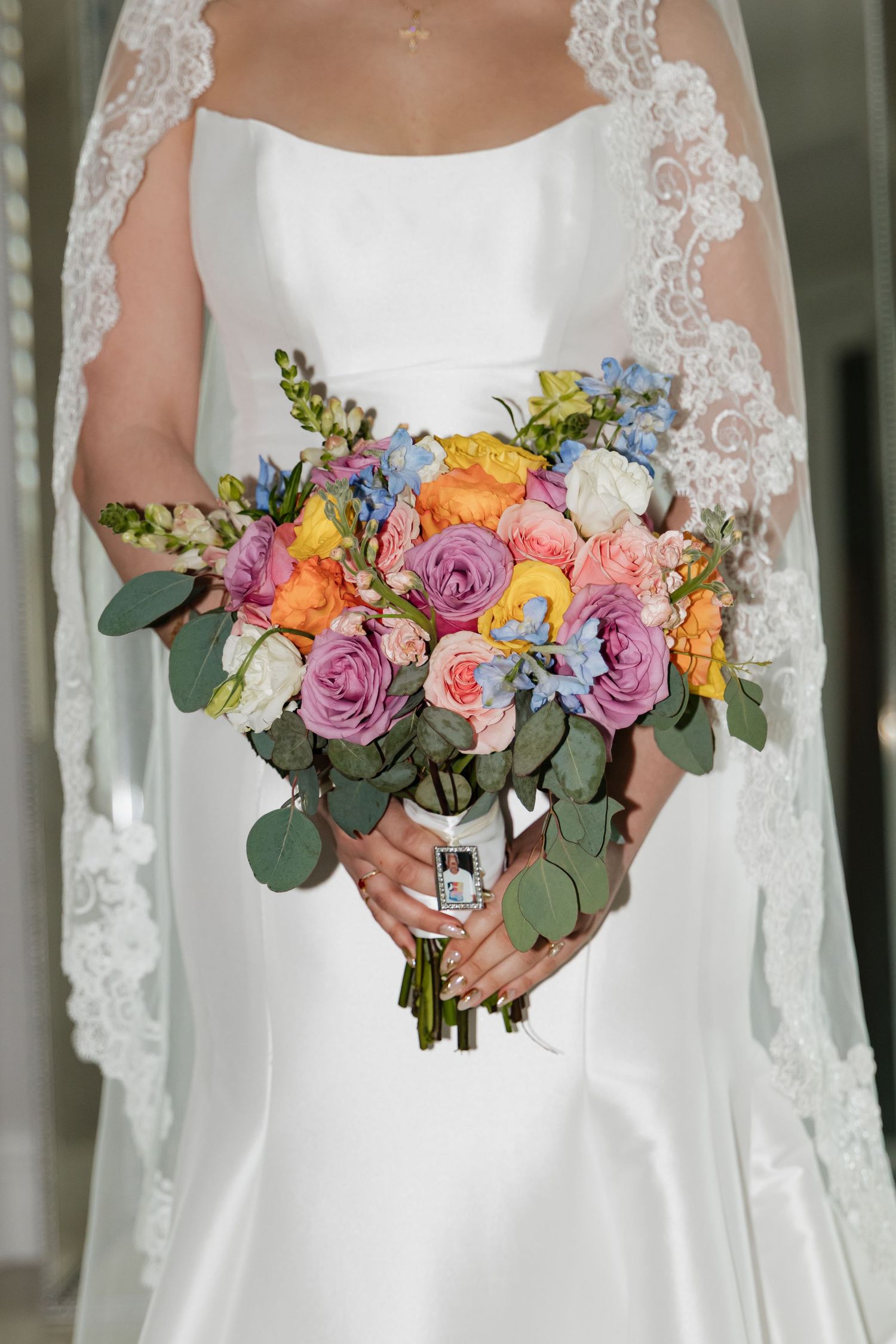Bride holding a colorful wedding bouquet with pink, orange, yellow, and blue roses in a white satin gown with lace veil.