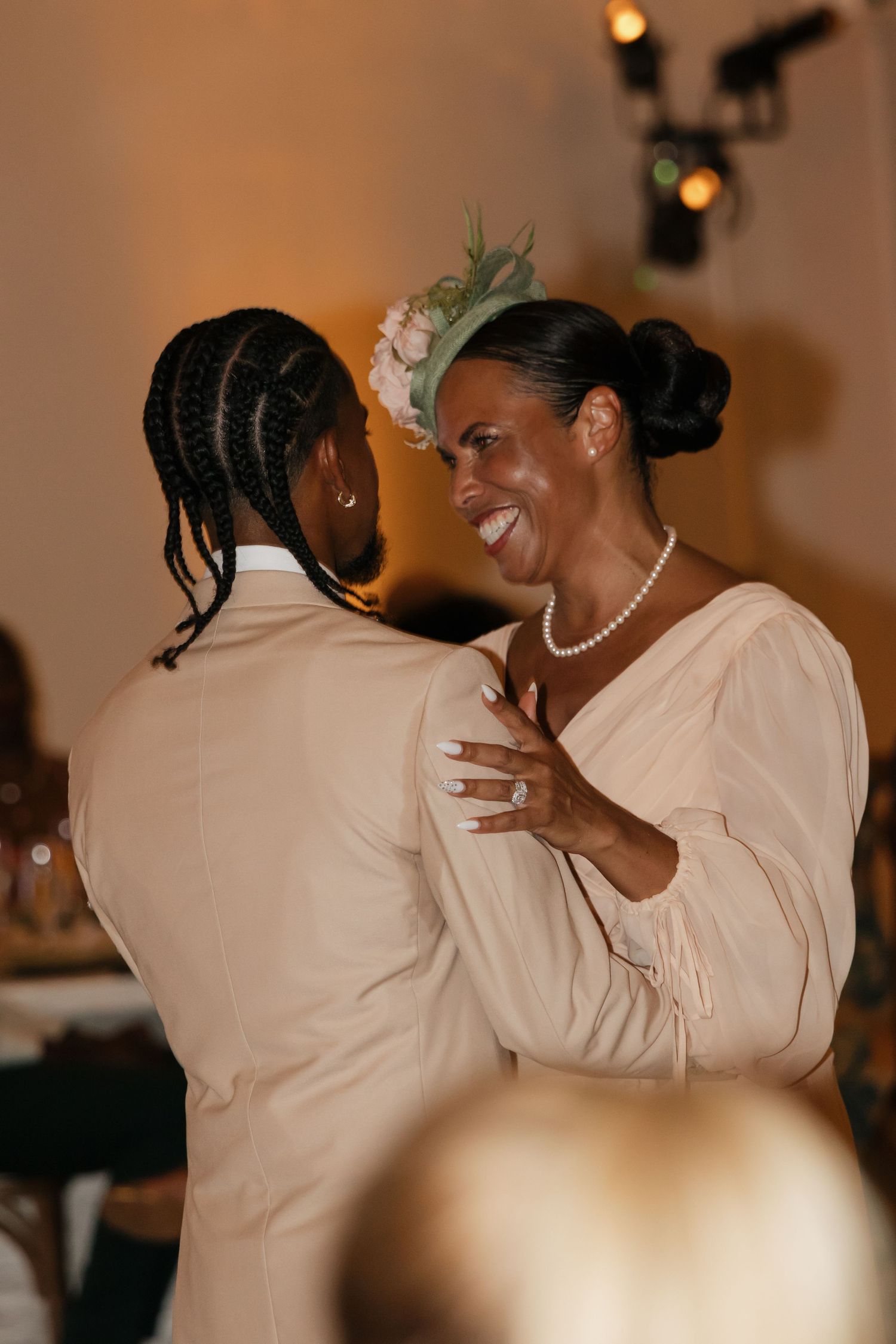 Groom shares joyful mother-son dance with woman in pearl necklace and floral fascinator hat.