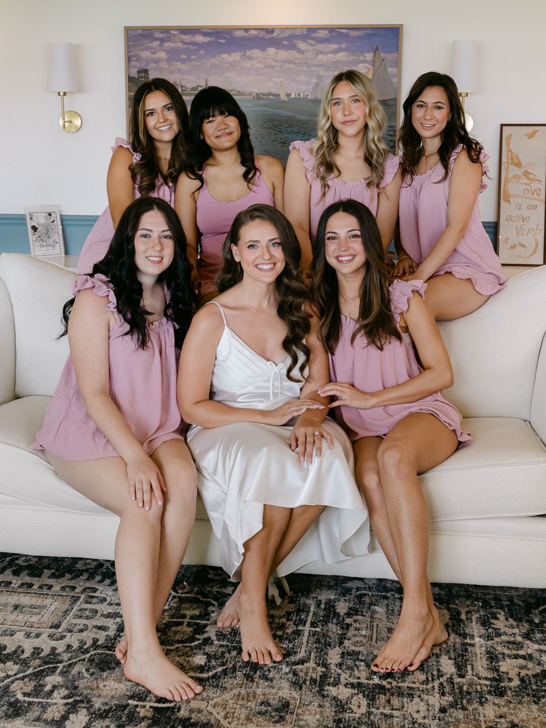 Bride in white gown poses with six bridesmaids in pink rompers on a white couch during wedding preparations.