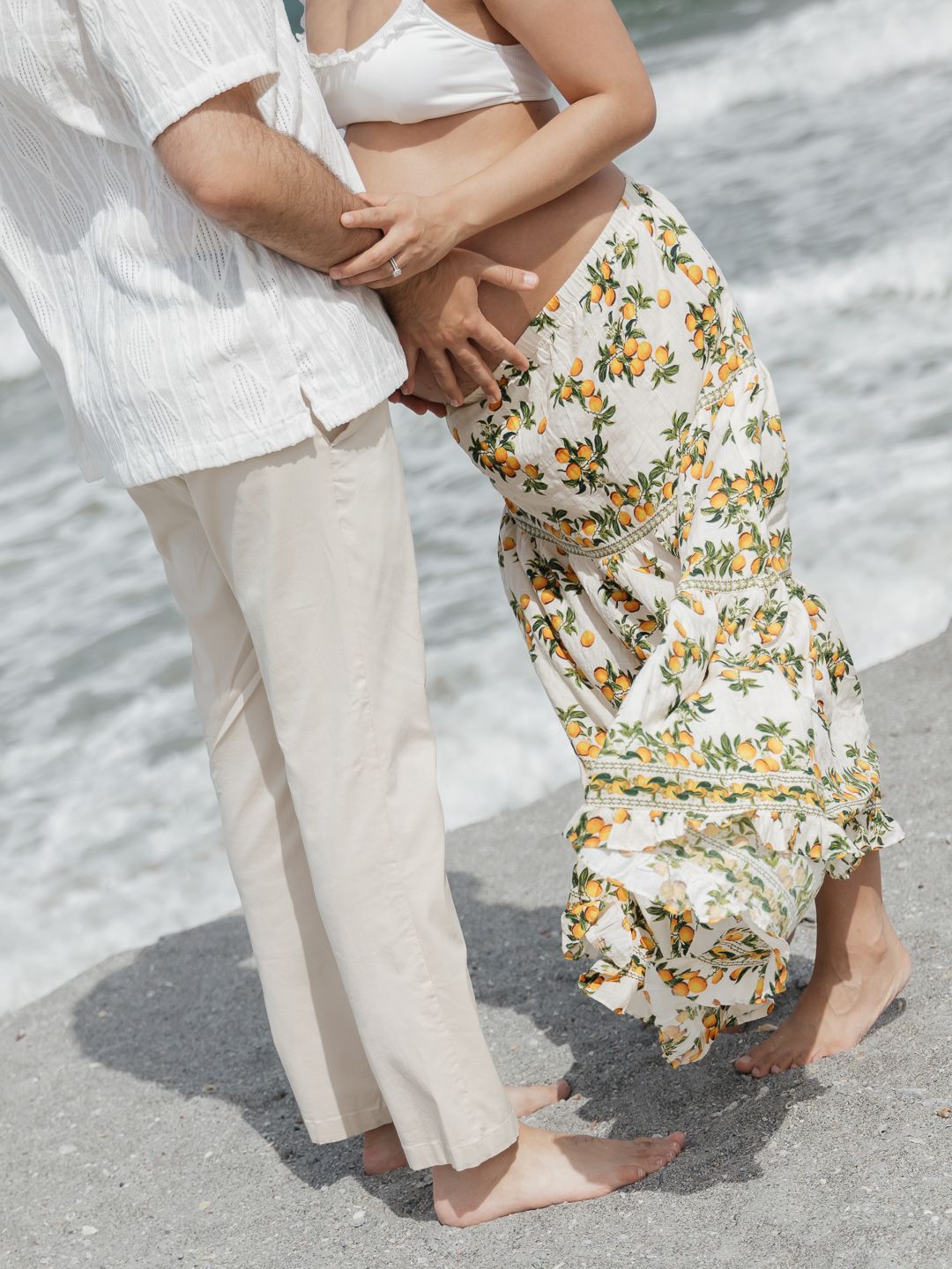 Couple embracing on beach, woman in floral maxi skirt and crop top, man in white linen outfit, barefoot on sand.