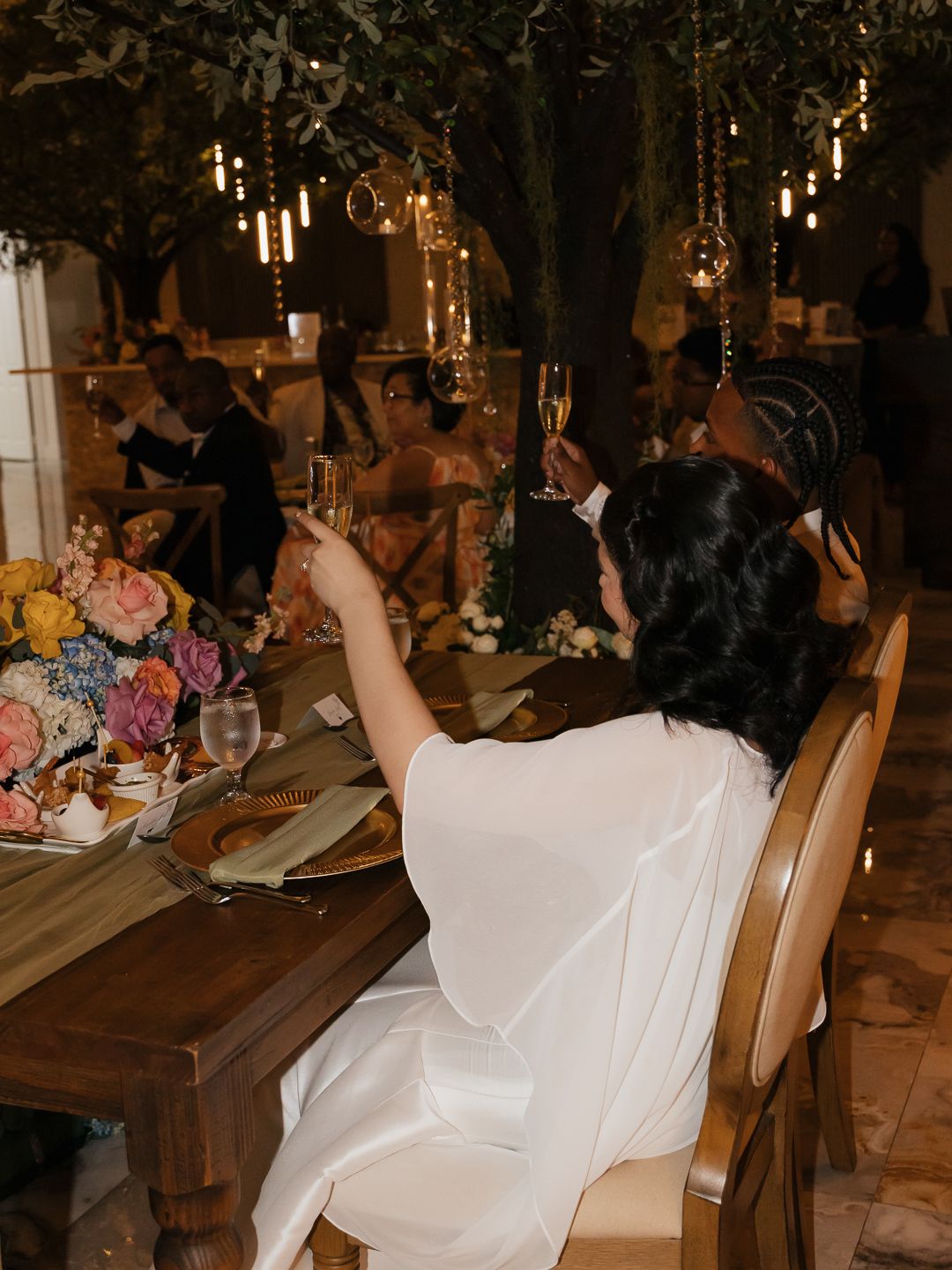Bride in white cape gown raises champagne toast at romantic candlelit wedding reception dinner.