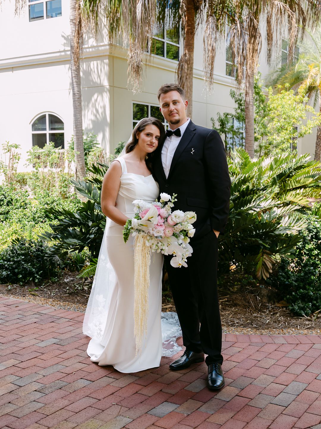 Bride and groom pose together in front of tropical venue, she in white gown, he in black tuxedo.