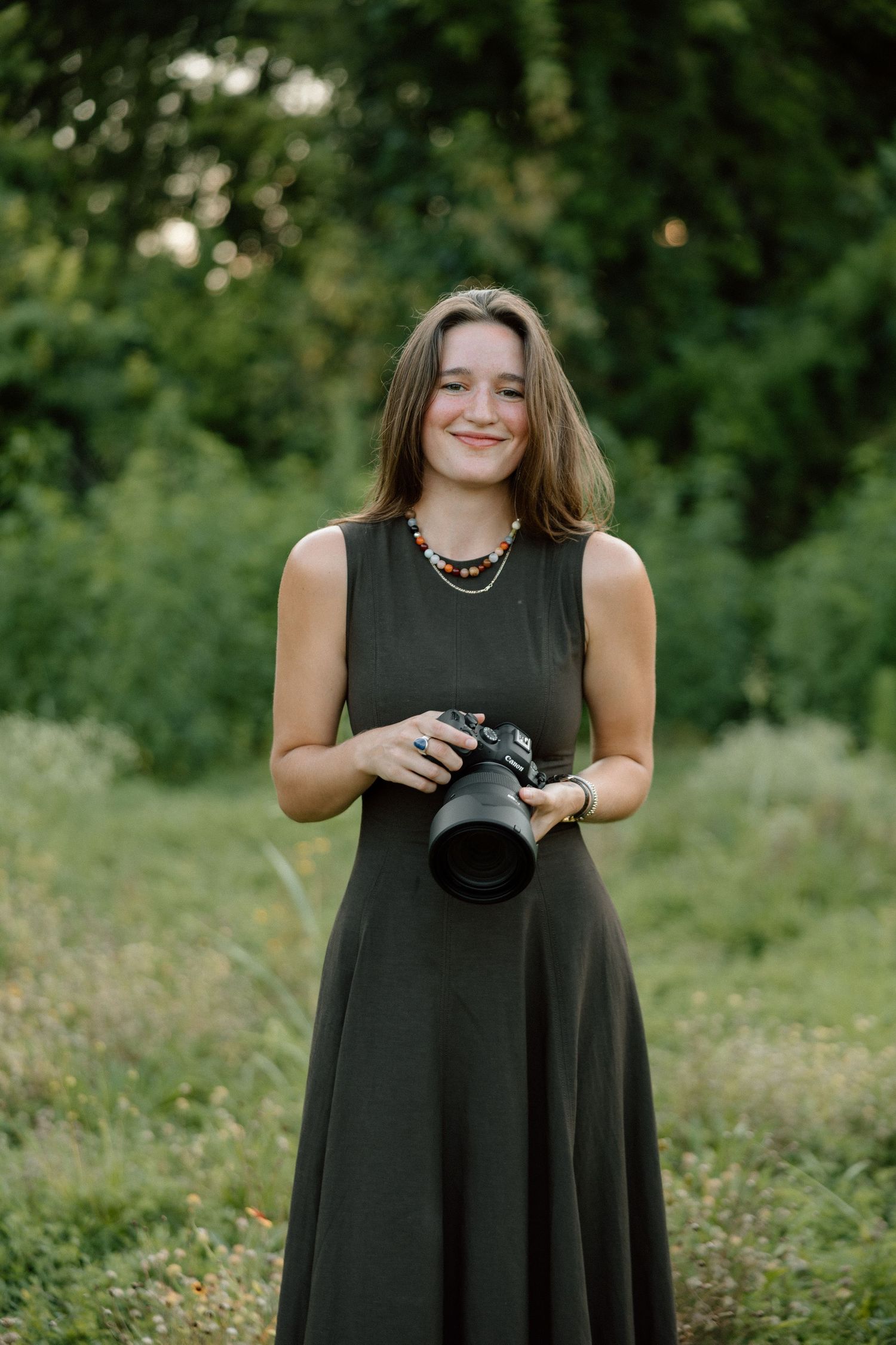 A photographer in a flowing black dress stands in a sunlit meadow holding a professional camera.