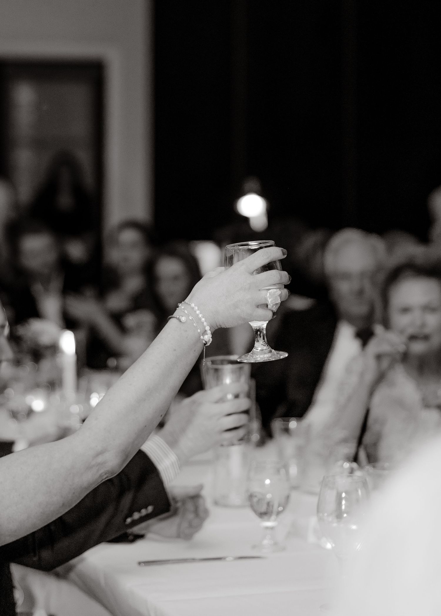 A black and white photograph shows multiple hands raised in a toast with wine glasses at a formal dining event.