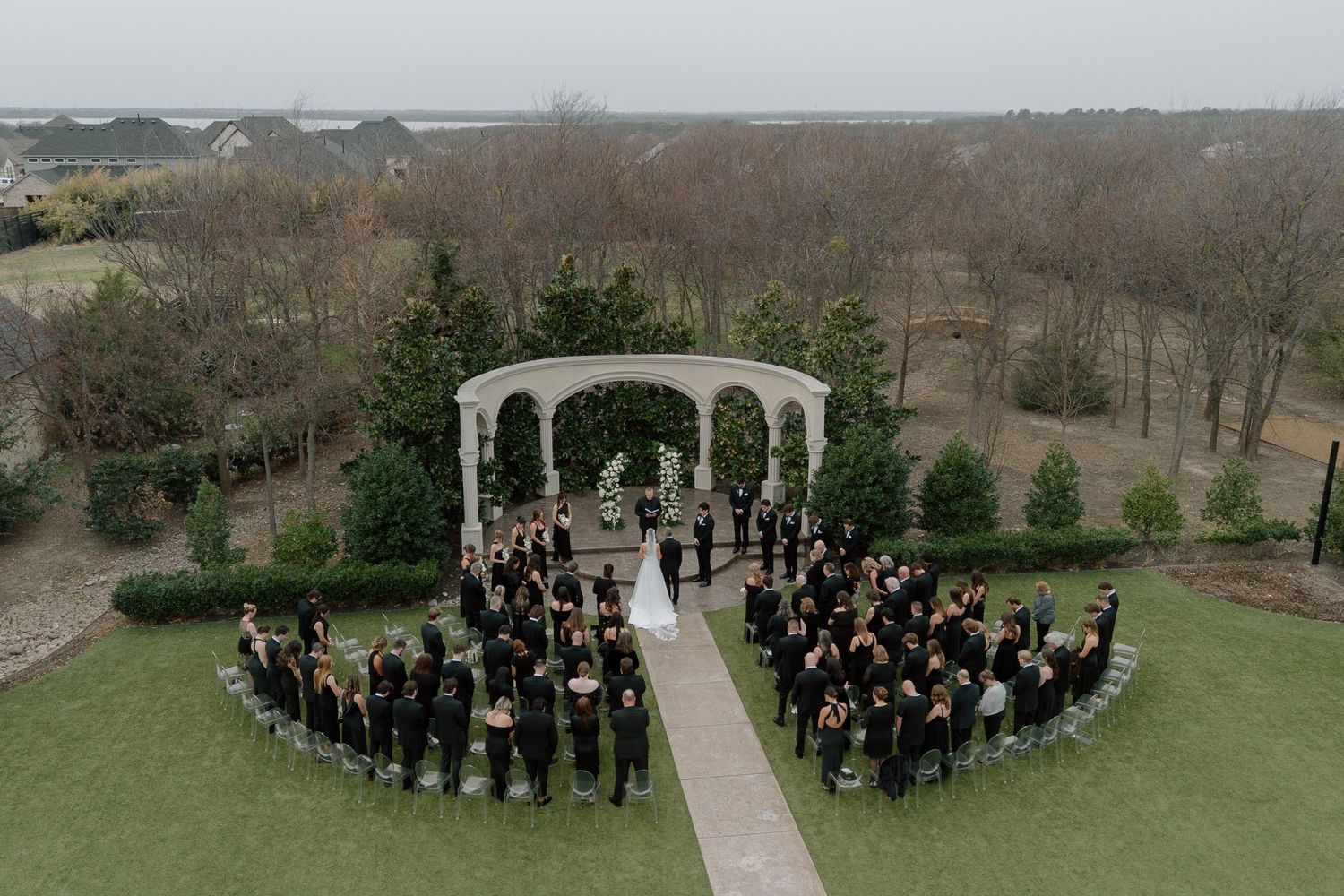 Aerial view of an elegant outdoor wedding ceremony under a white gazebo surrounded by guests in black attire on lush green grounds.