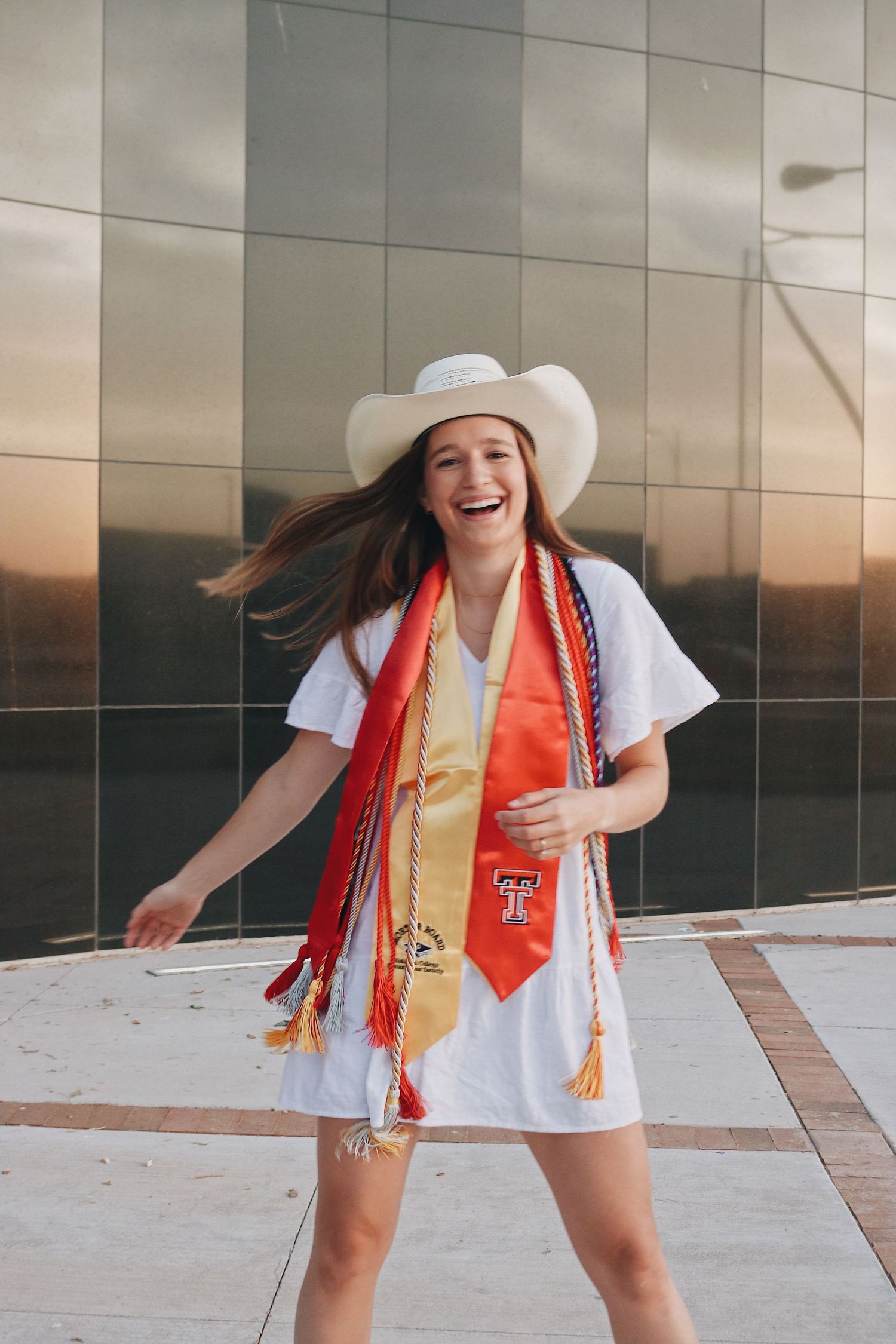 Graduate in white dress and orange stole poses playfully in front of reflective building wall during sunset photoshoot.