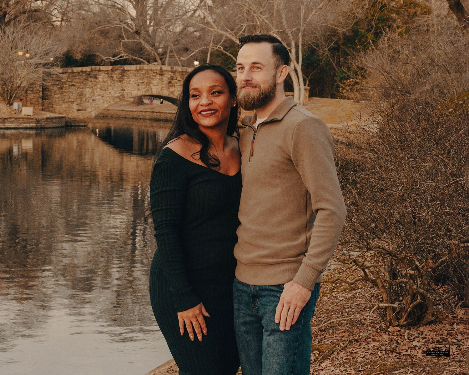 Couple posing together by a scenic pond with a stone bridge in a park during winter.