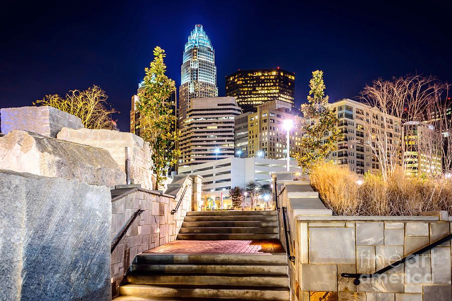 Charlotte NC skyline at night with illuminated skyscrapers, stone steps, and trees in Romare Bearden Park.