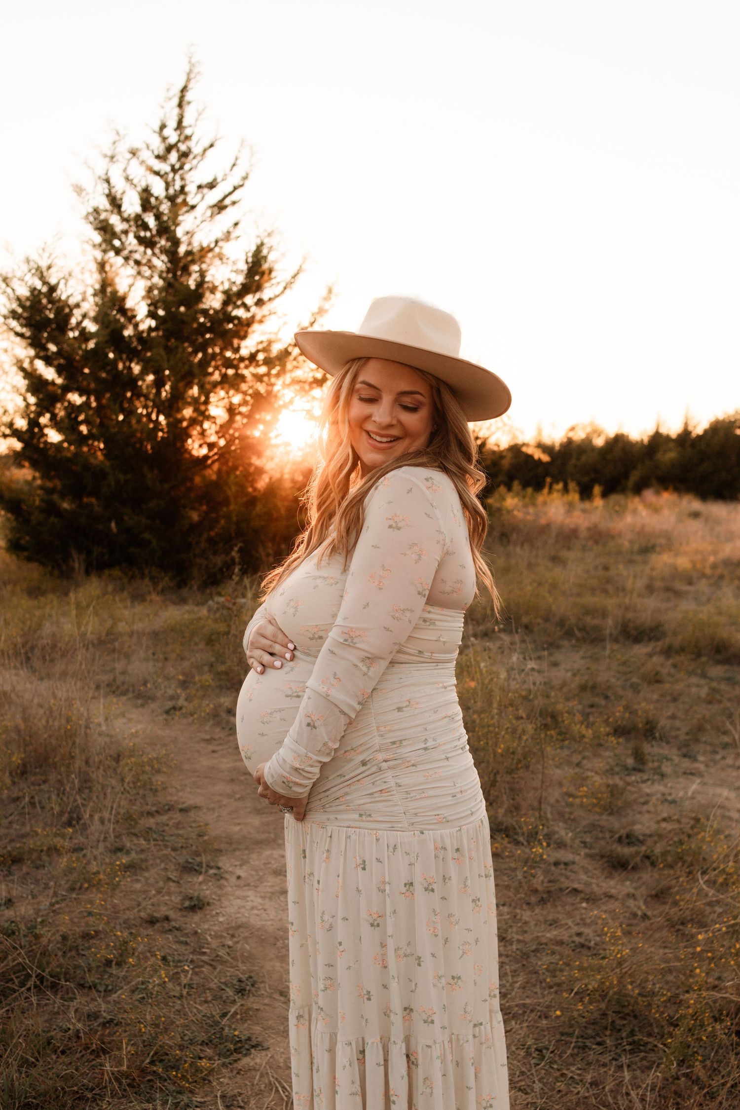 A pregnant woman in a white dress and hat poses during golden hour sunset.