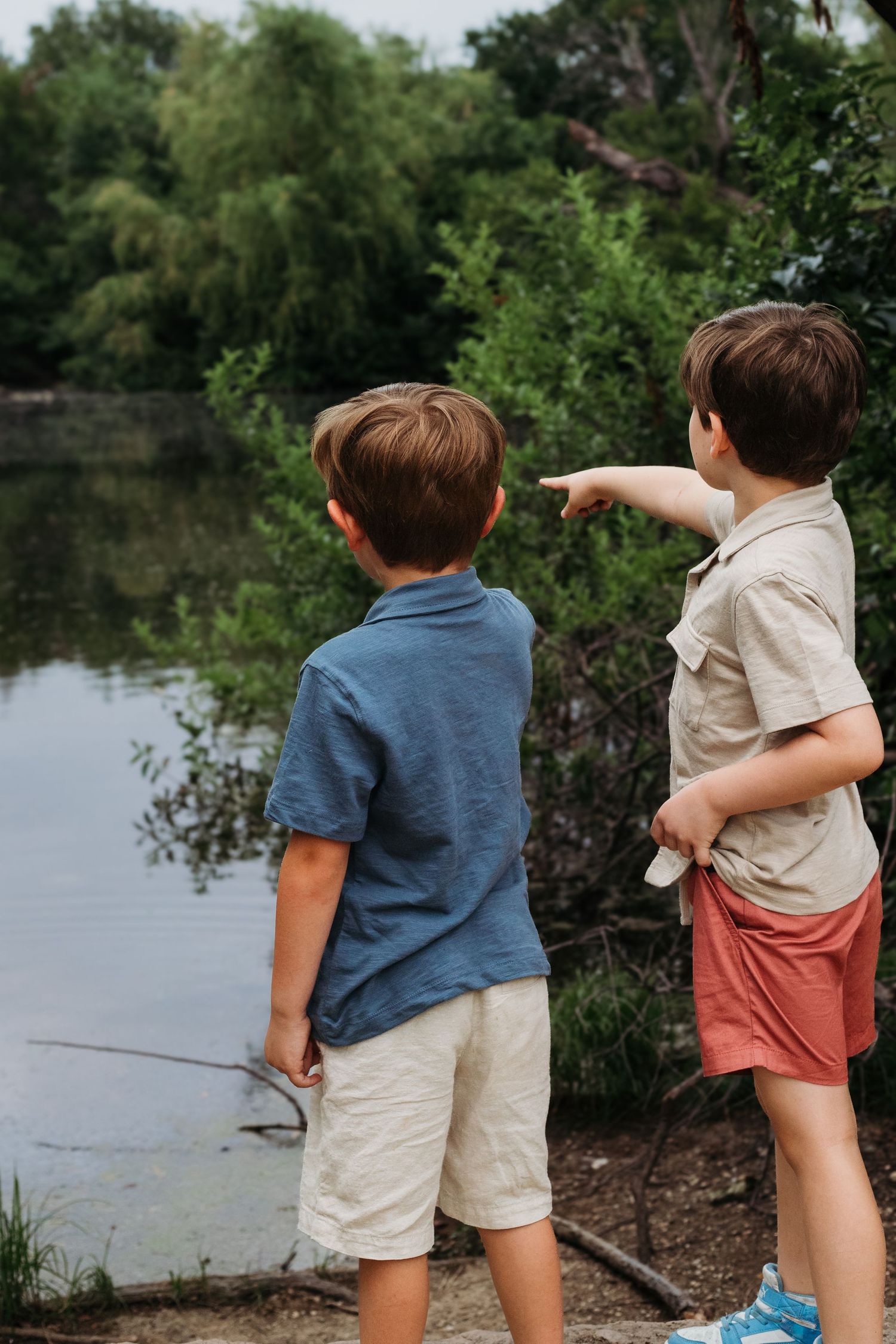 Two children point at something while standing by a peaceful pond surrounded by trees.