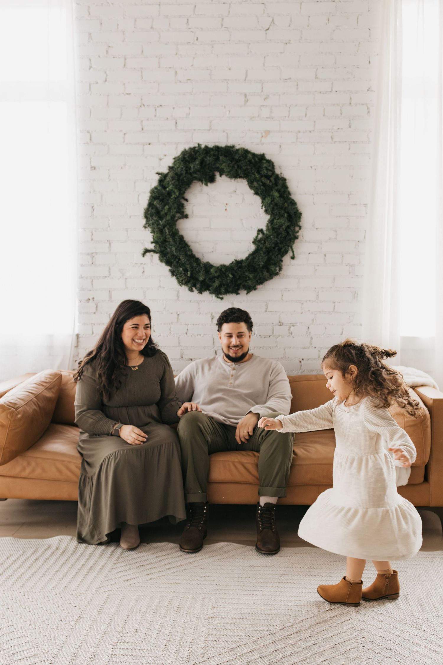 Family sits on leather couch beneath wreath while child dances in white dress.