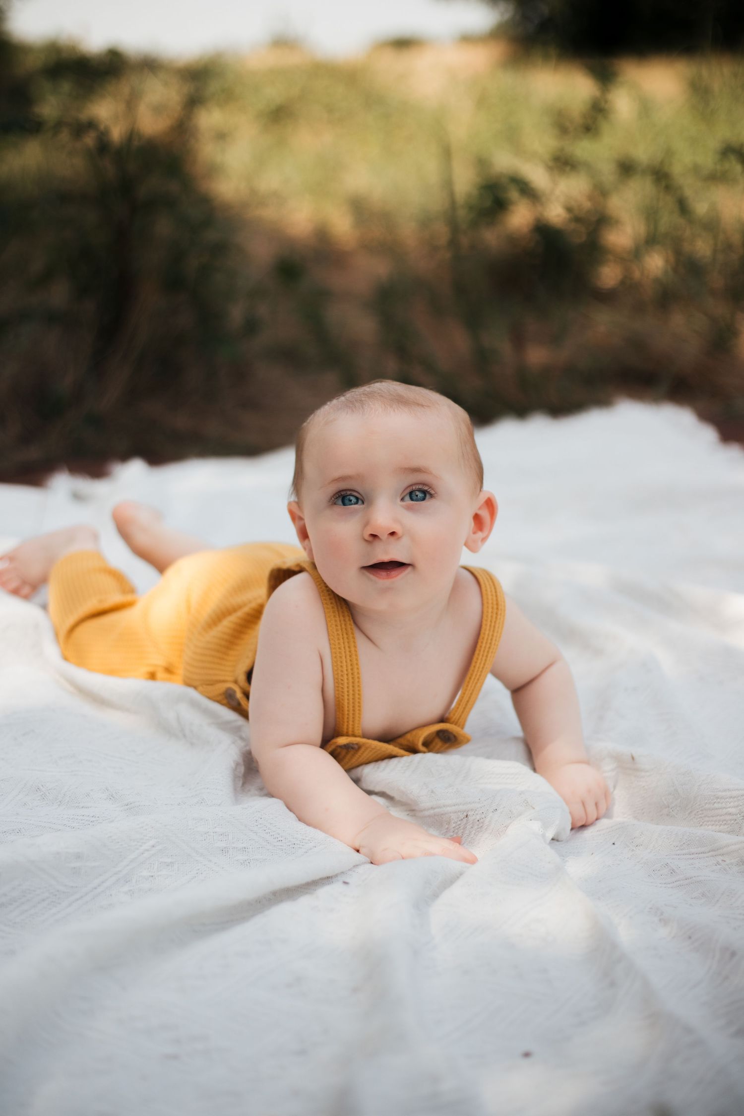 Baby in yellow romper lying on white blanket outdoors during golden hour.