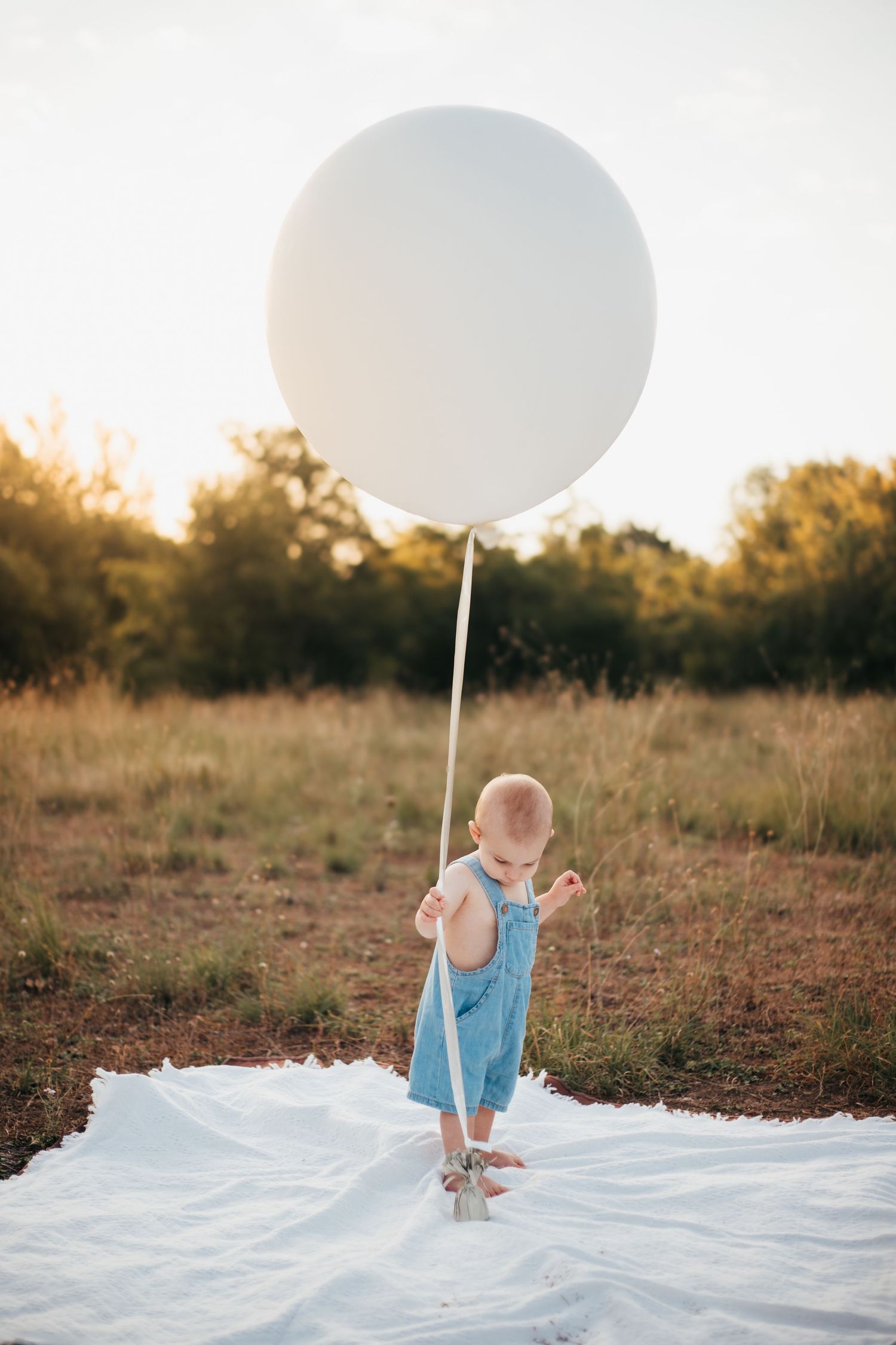 Small child stands on white blanket holding large white balloon in golden sunset field.