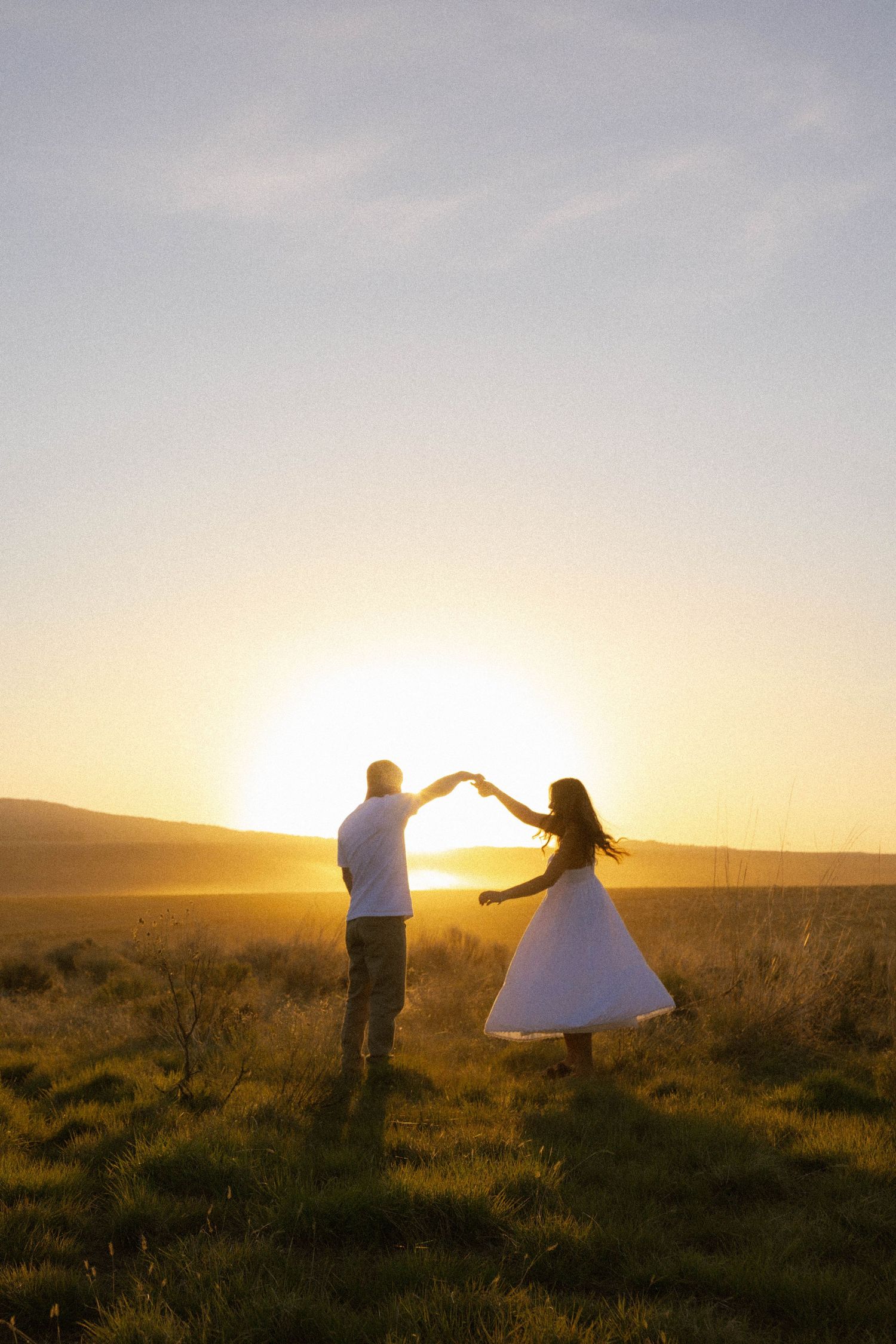 Couple dancing at golden sunset in open field, woman in white dress twirling under raised hands.