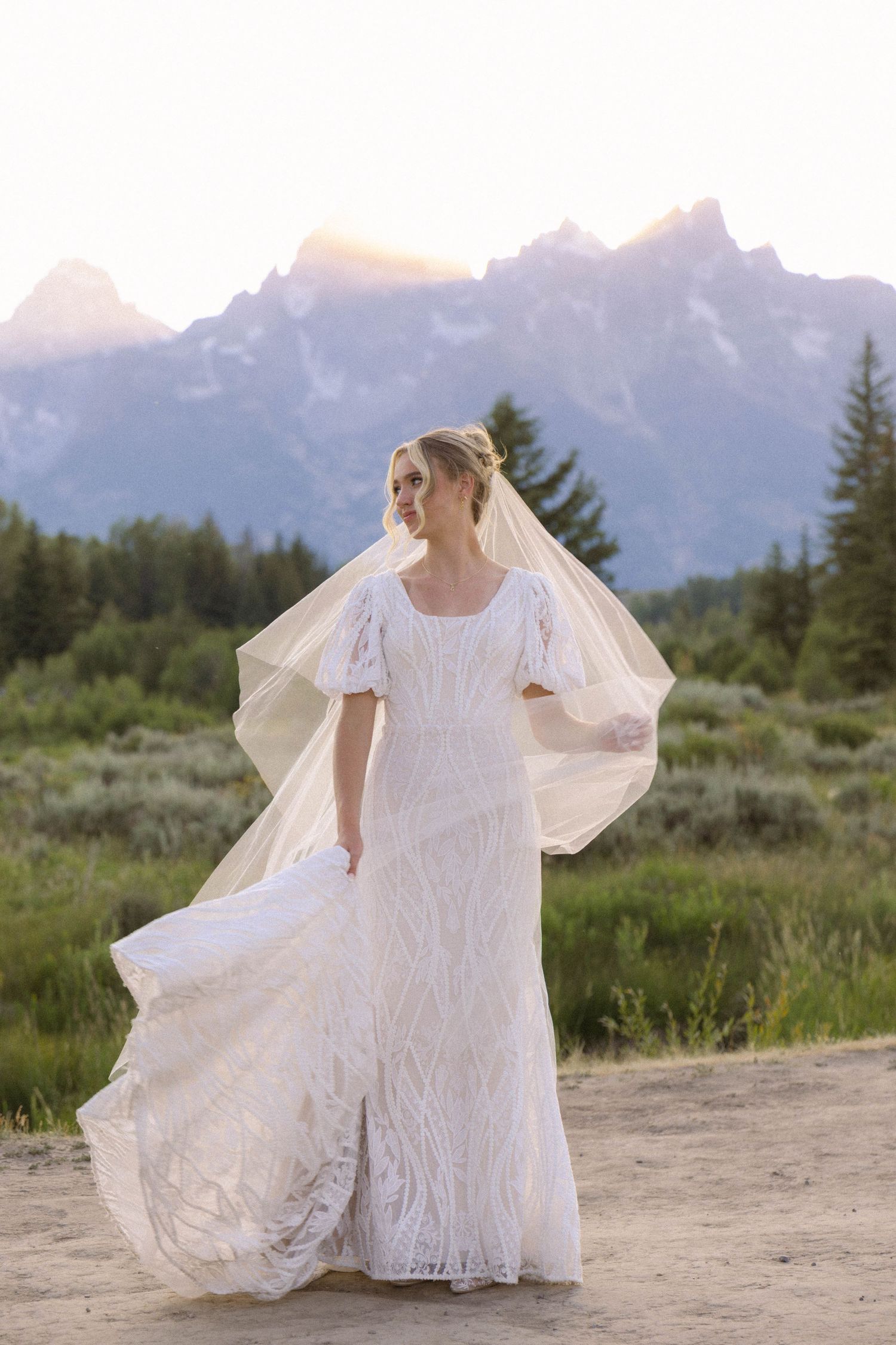 Bride in white lace gown and flowing veil poses before Grand Teton mountains at sunset.