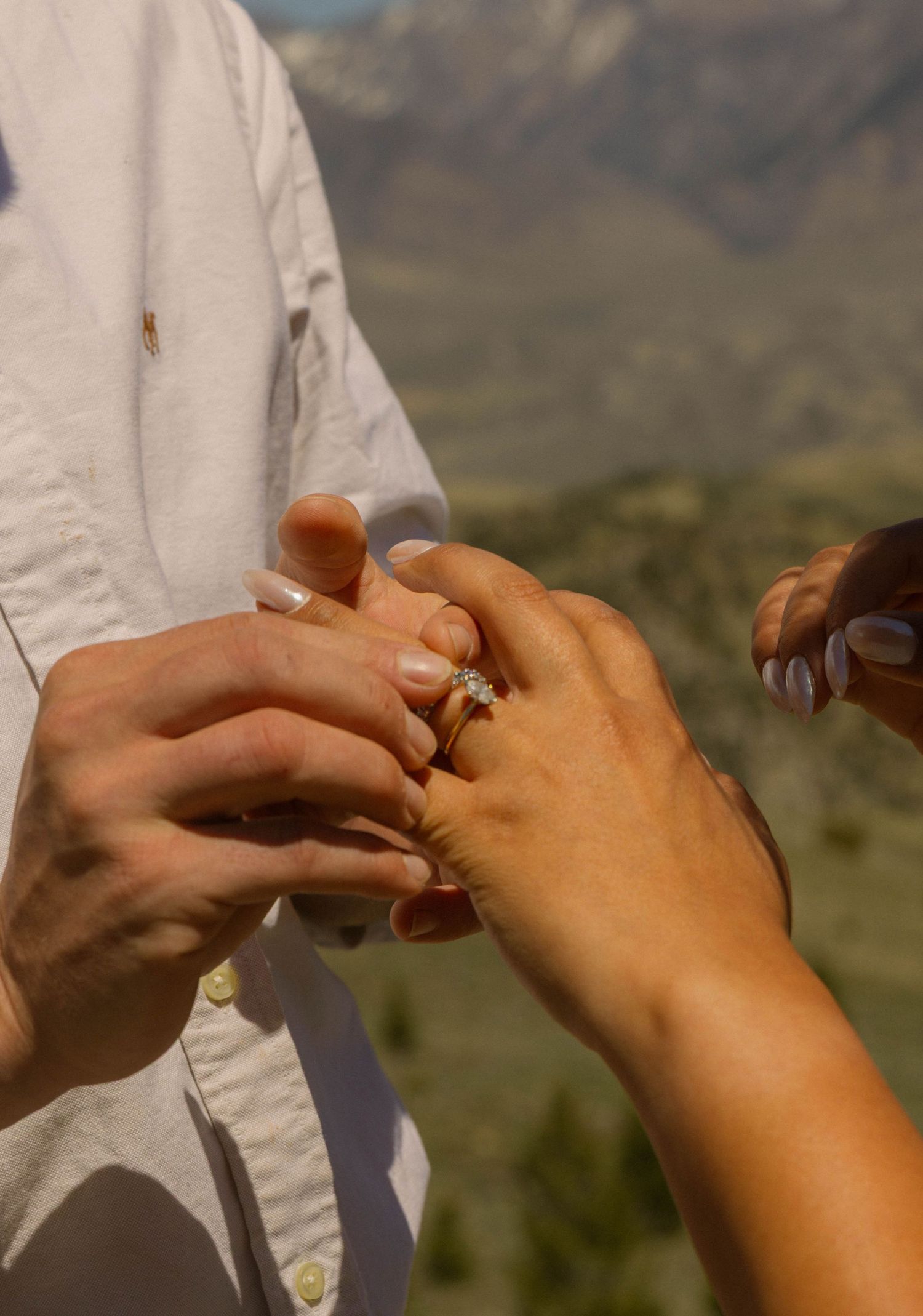 Person placing a diamond engagement ring on partner's finger with scenic mountain backdrop during outdoor proposal.