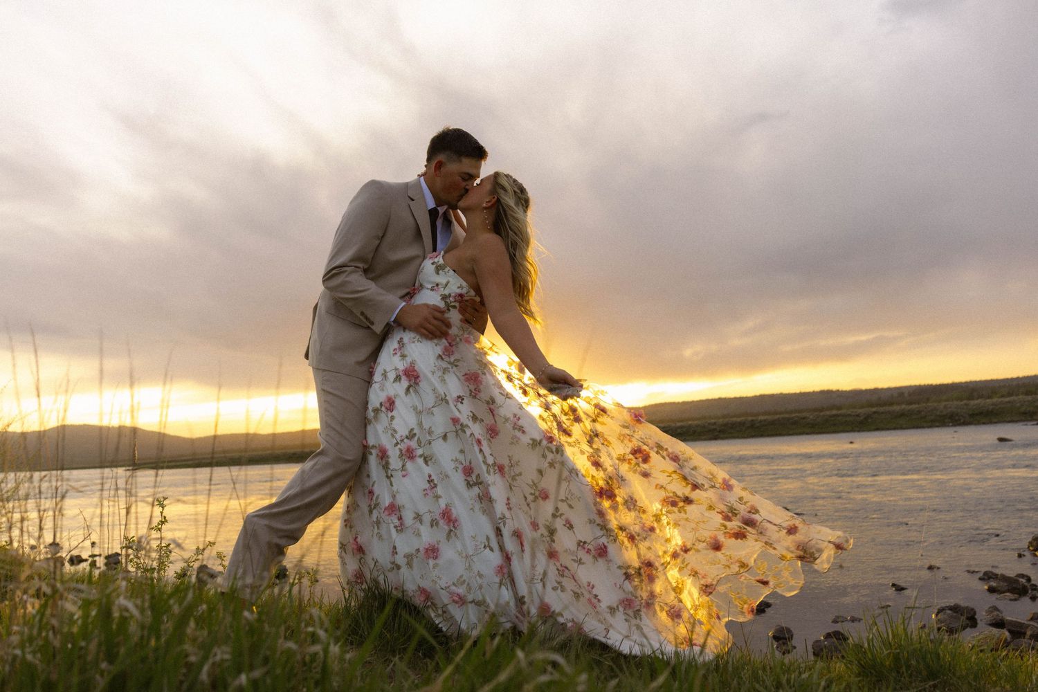 Couple sharing a romantic kiss at sunset by a lake, bride in a floral gown, groom in a tan suit.