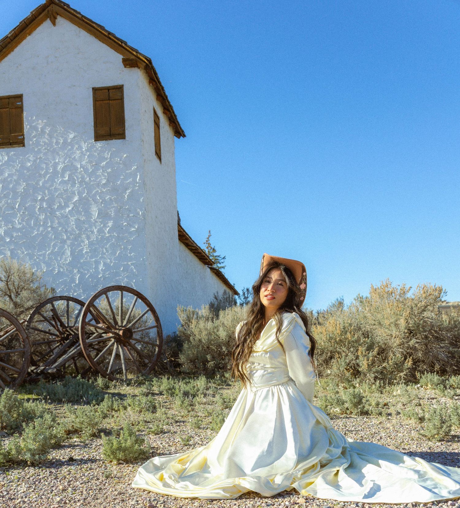 Woman in vintage white gown kneeling near rustic wagon wheel by white adobe building under clear blue sky.